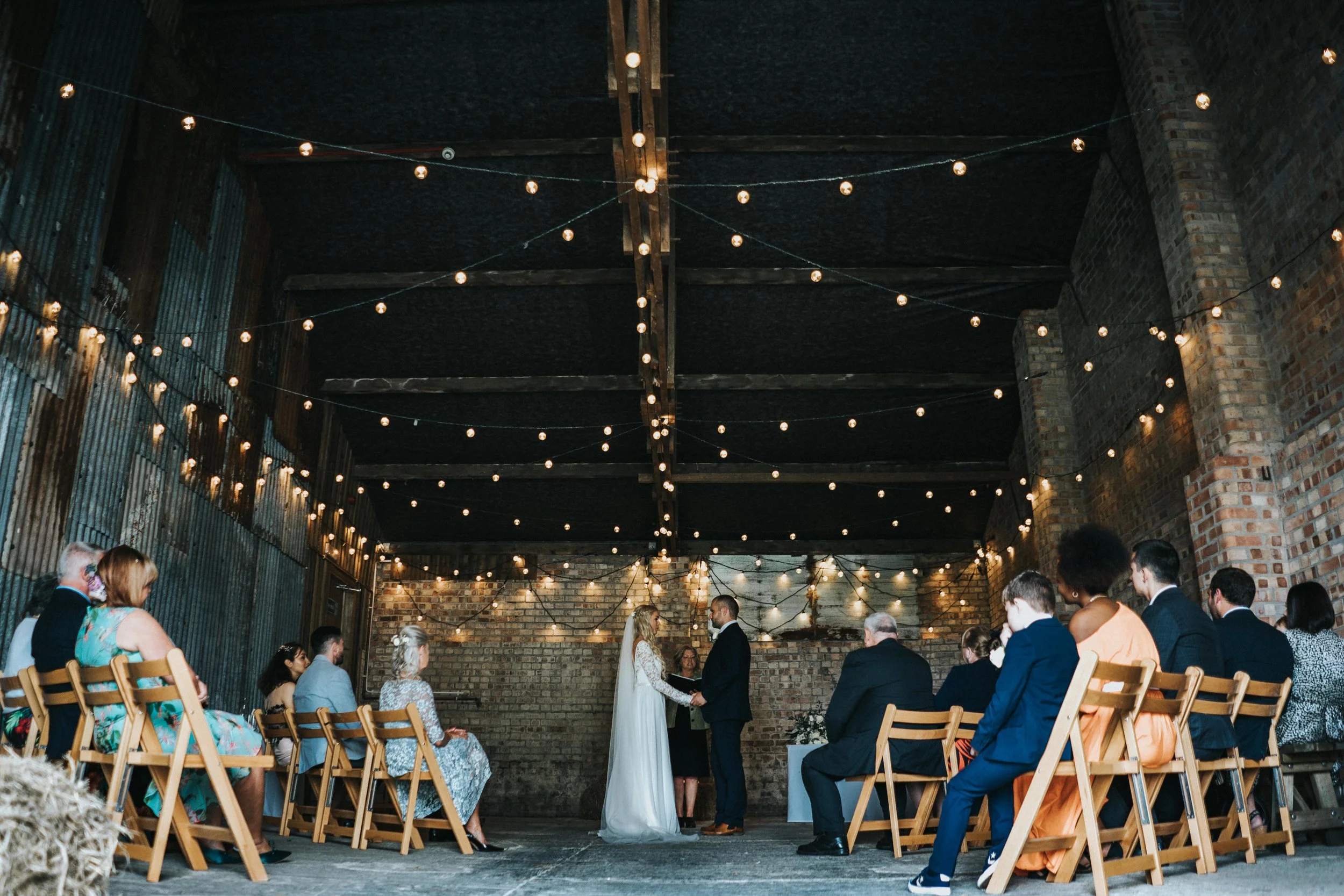 A wedding ceremony with the bride and groom holding hands at the center, facing each other, in an industrial-style venue with exposed brick walls and wooden beams. Guests are seated on both sides in wooden chairs under string lights hanging from the 