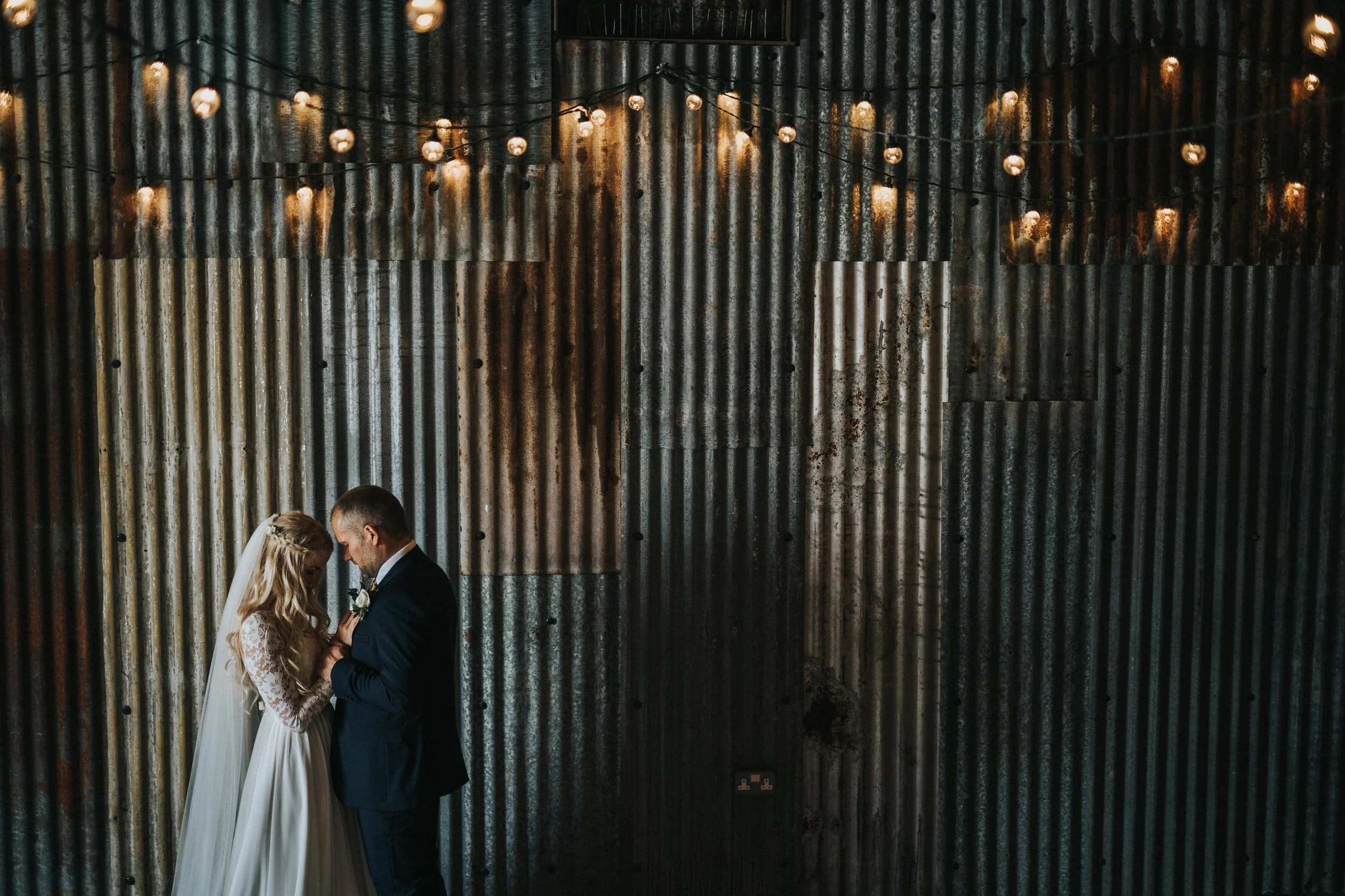 A bride and groom standing close together against a corrugated metal wall with string lights overhead.