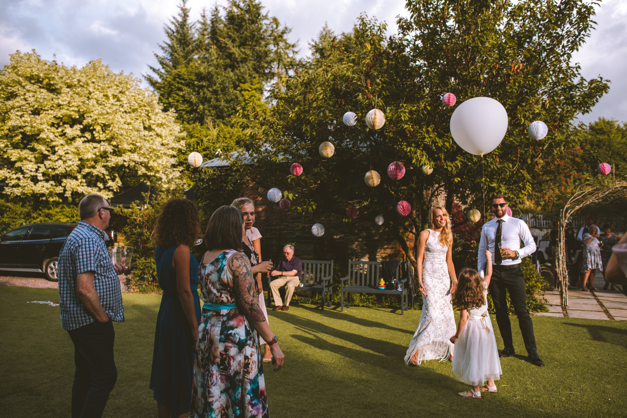 People celebrating outdoors. A woman in a white lace dress and a man in a black suit hold hands with a young girl in a white dress. Other guests stand nearby, with one man in sunglasses and a white shirt, holding a drink. The background has trees wit