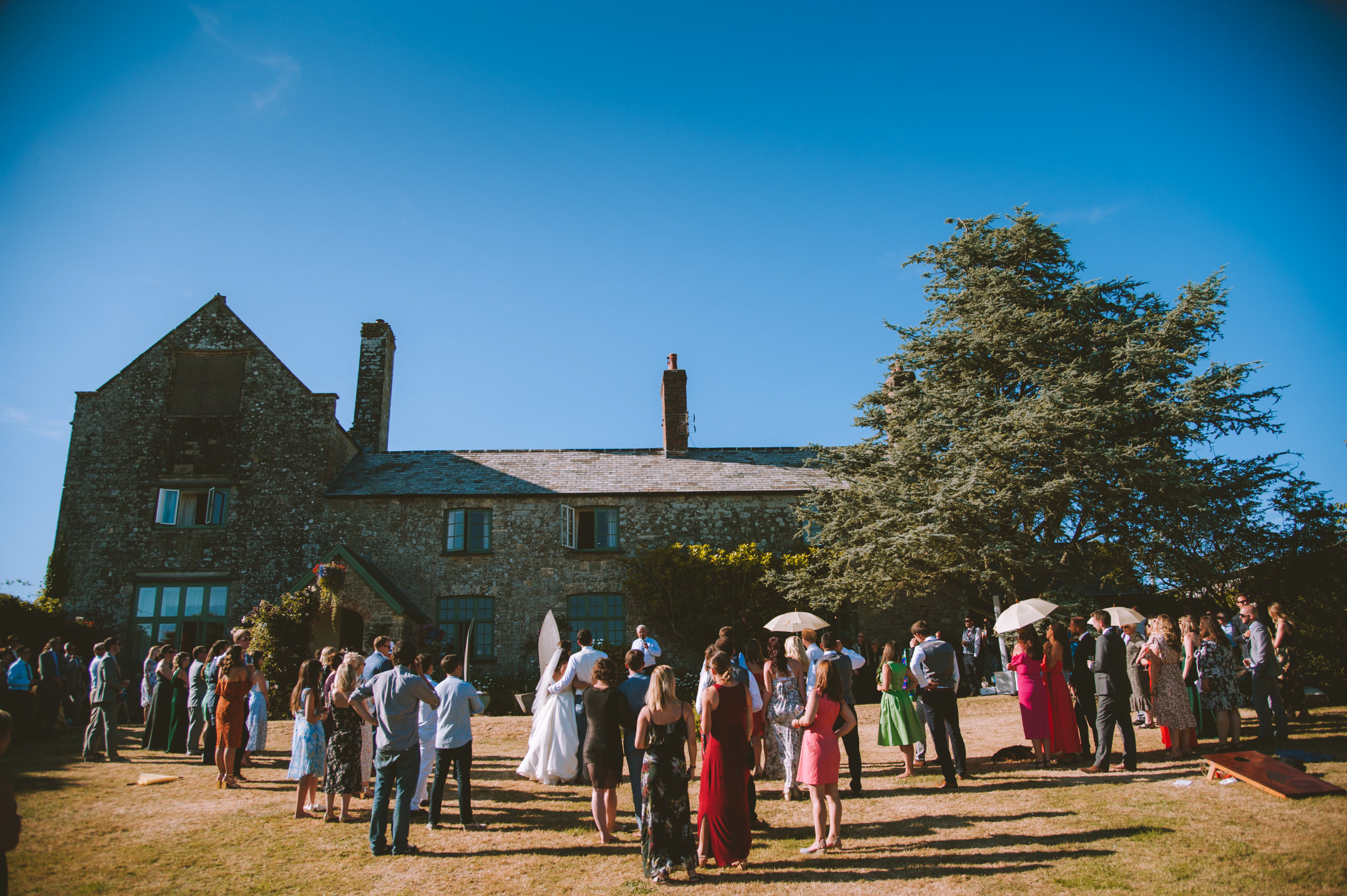Wedding reception outdoors with guests holding umbrellas, in front of a historic stone building on a sunny day.