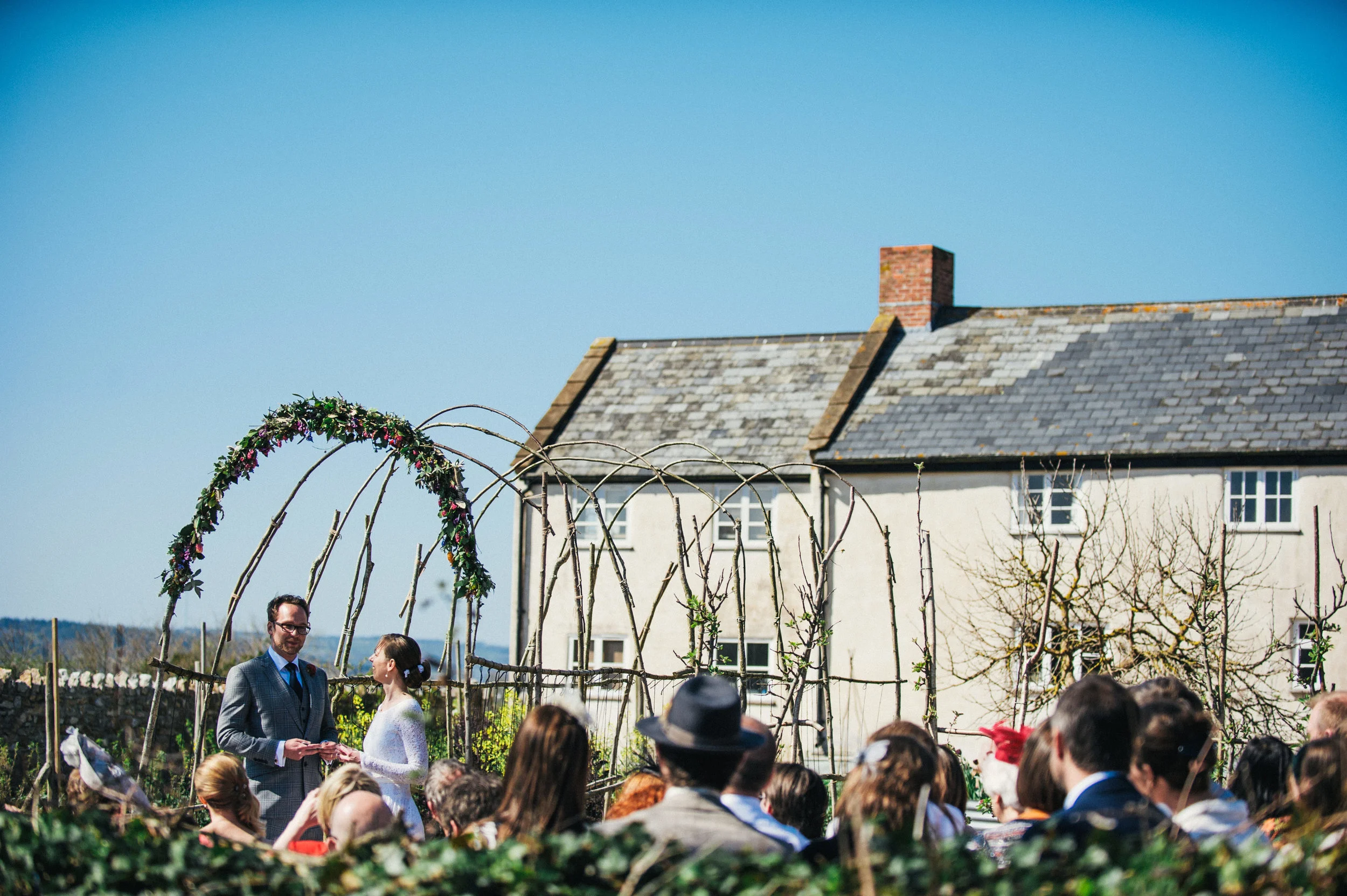 Wedding ceremony outdoors with a couple standing under a floral arch, surrounded by guests, in front of a rustic house on a sunny day.