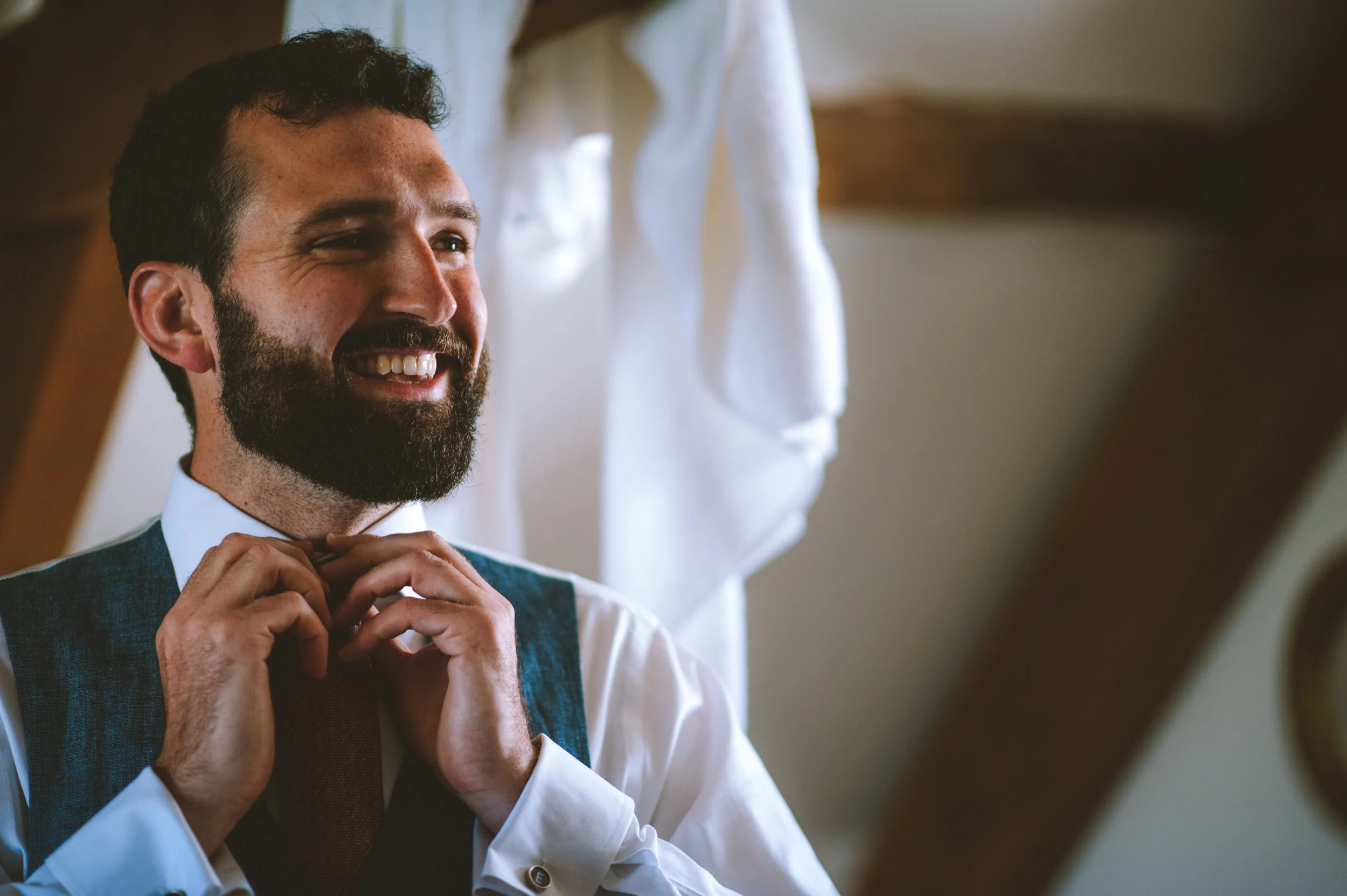 A man with a beard smiling as he adjusts his bow tie, wearing a white shirt and vest, in a warmly lit room.