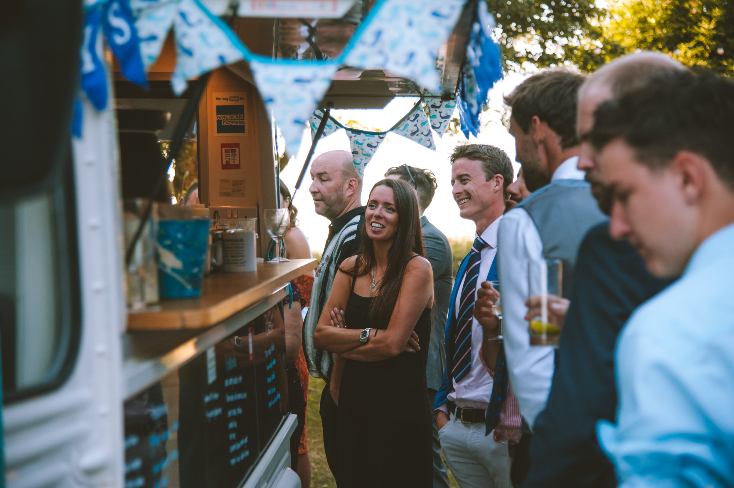 People gathered at an outdoor event, standing in line at a food or drink stand decorated with blue and white bunting.