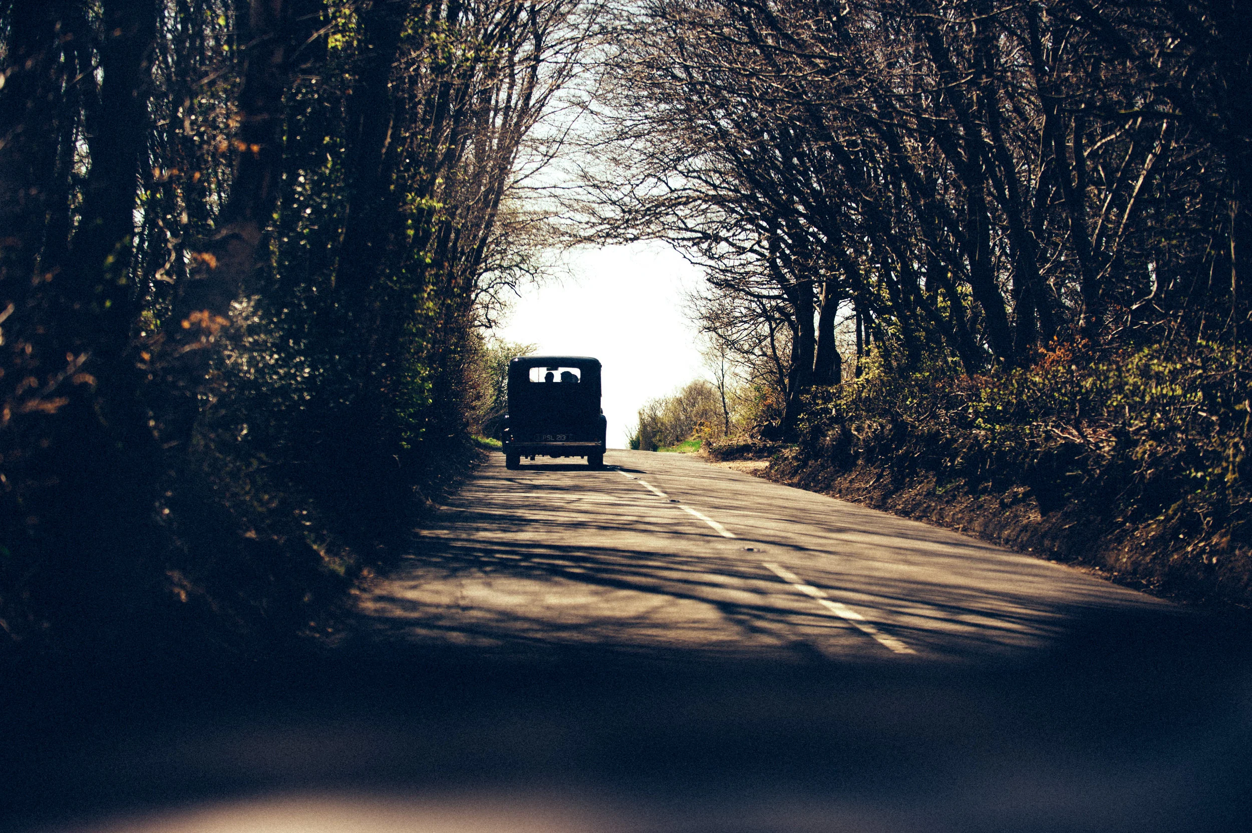 A black vintage van driving down a two-lane road surrounded by leafless trees with shadows on the road during daytime.
