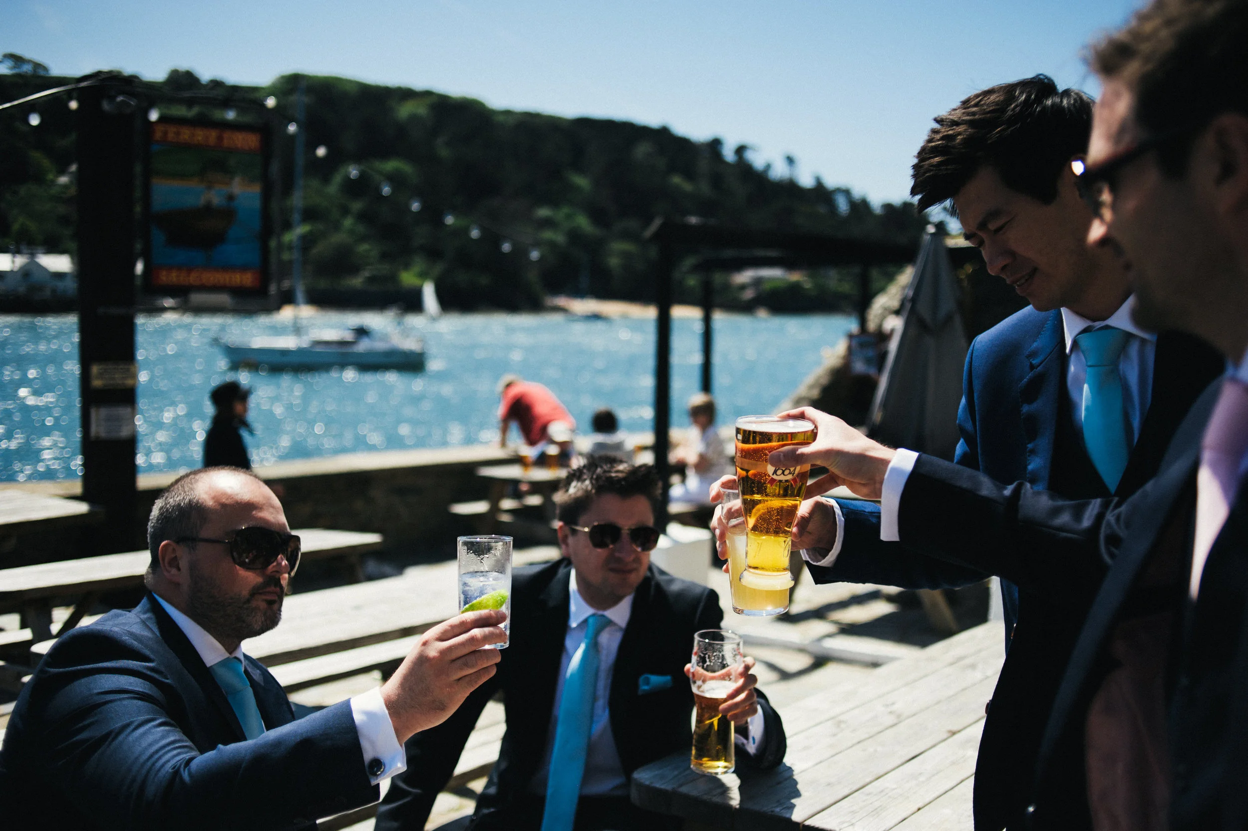 Four men in suits are sitting outdoors by a waterfront, with two holding glasses of beer, and one holding a cocktail with lime, during daytime with boats on the water in the background.