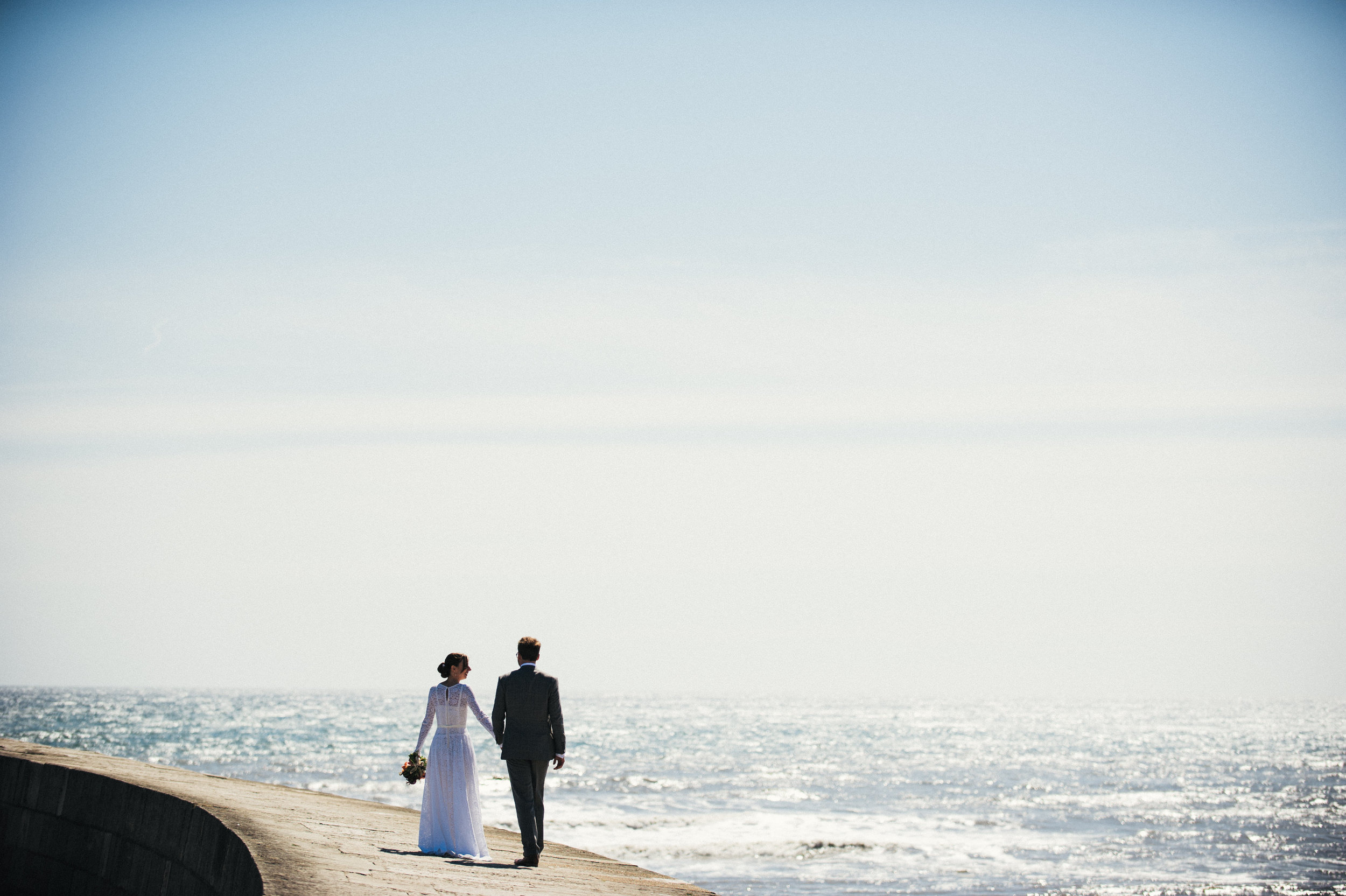 A bride and groom holding hands walk along a concrete walkway by the ocean, with the bride holding a bouquet.