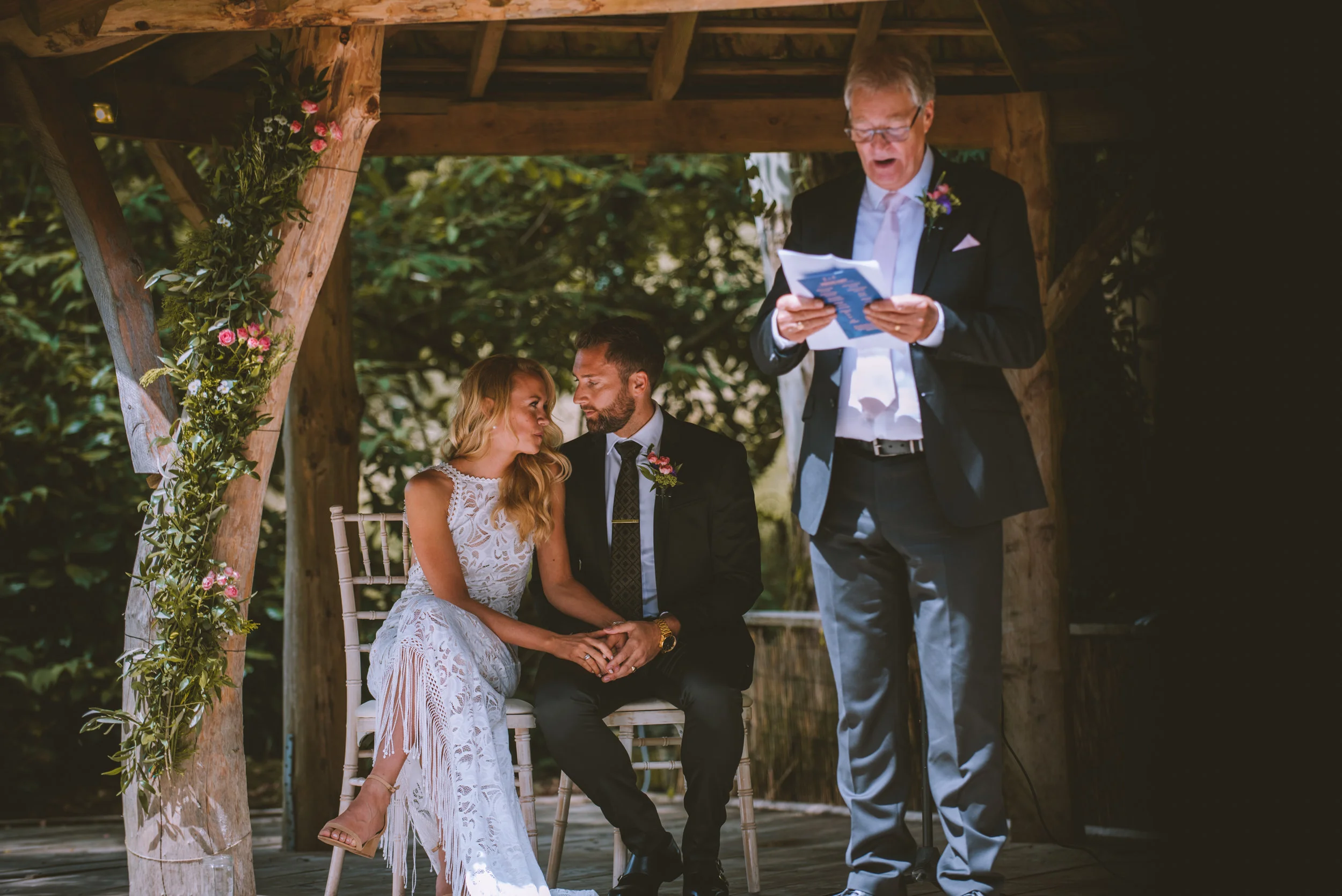 A couple sitting under a wooden wedding canopy, holding hands, with a man reading vows or a speech during an outdoor wedding ceremony.