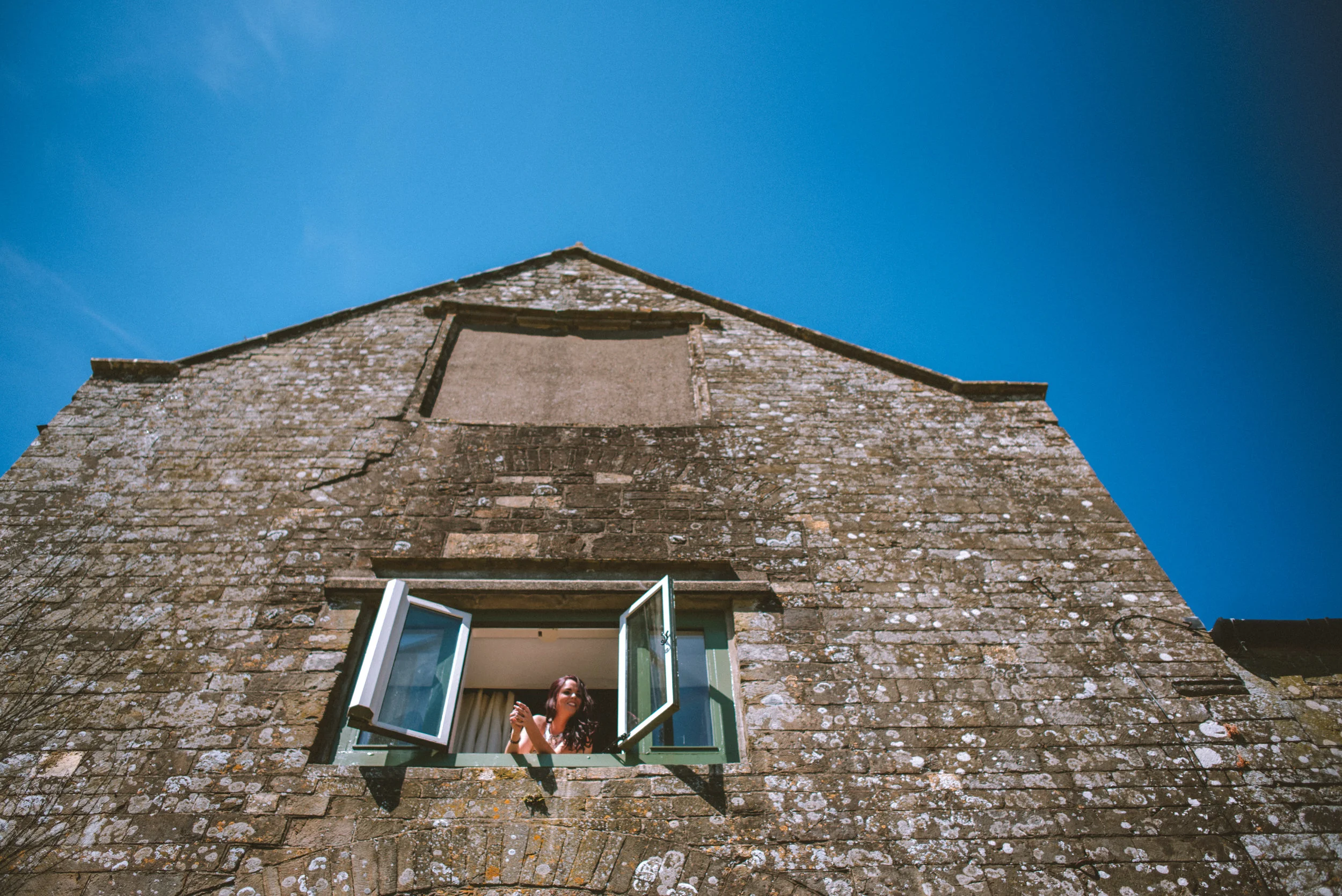 Woman smiling and looking out of an open window in an old brick building under a clear blue sky.