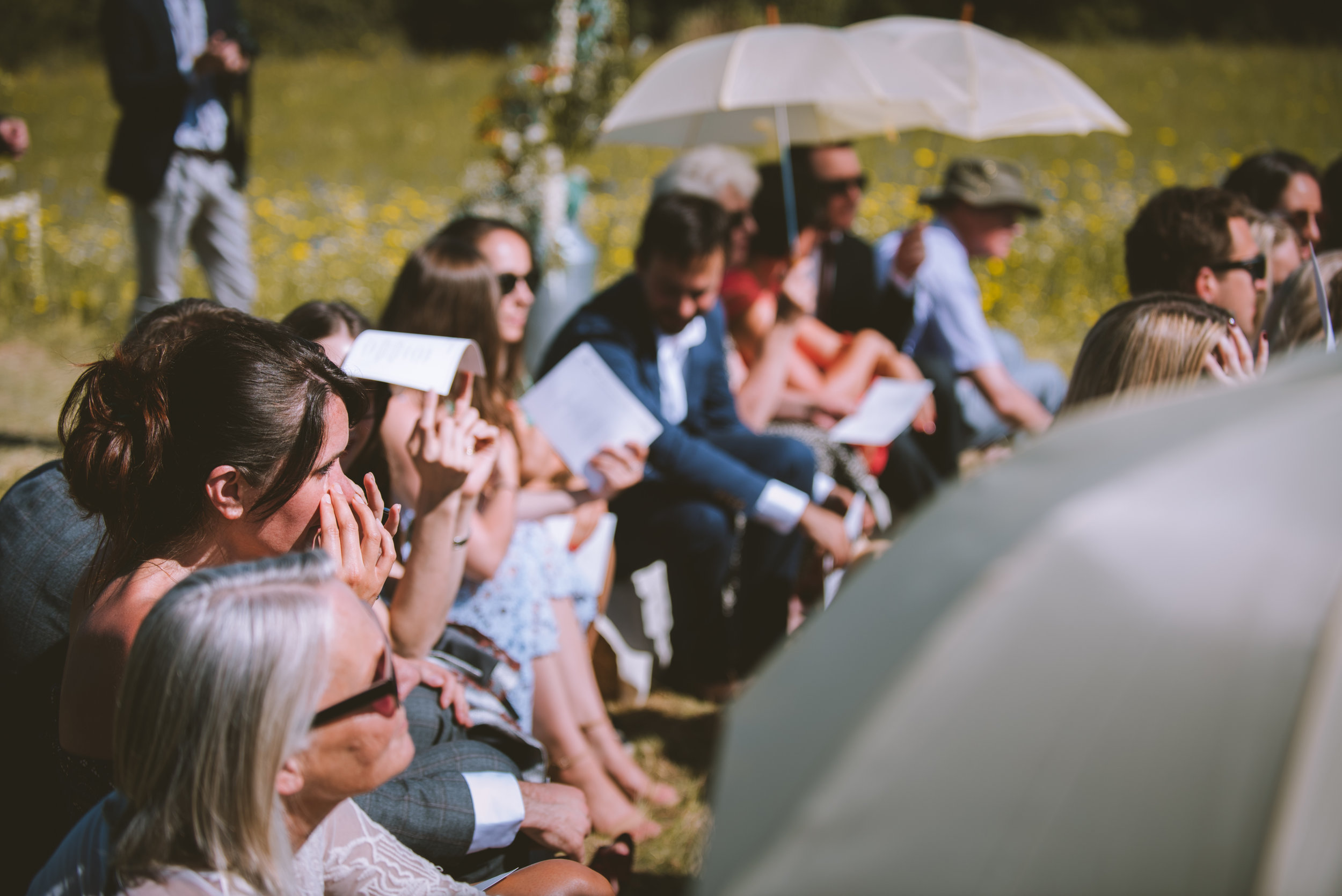 People attending an outdoor event, some with umbrellas, sitting on grass, many reading papers or programs.