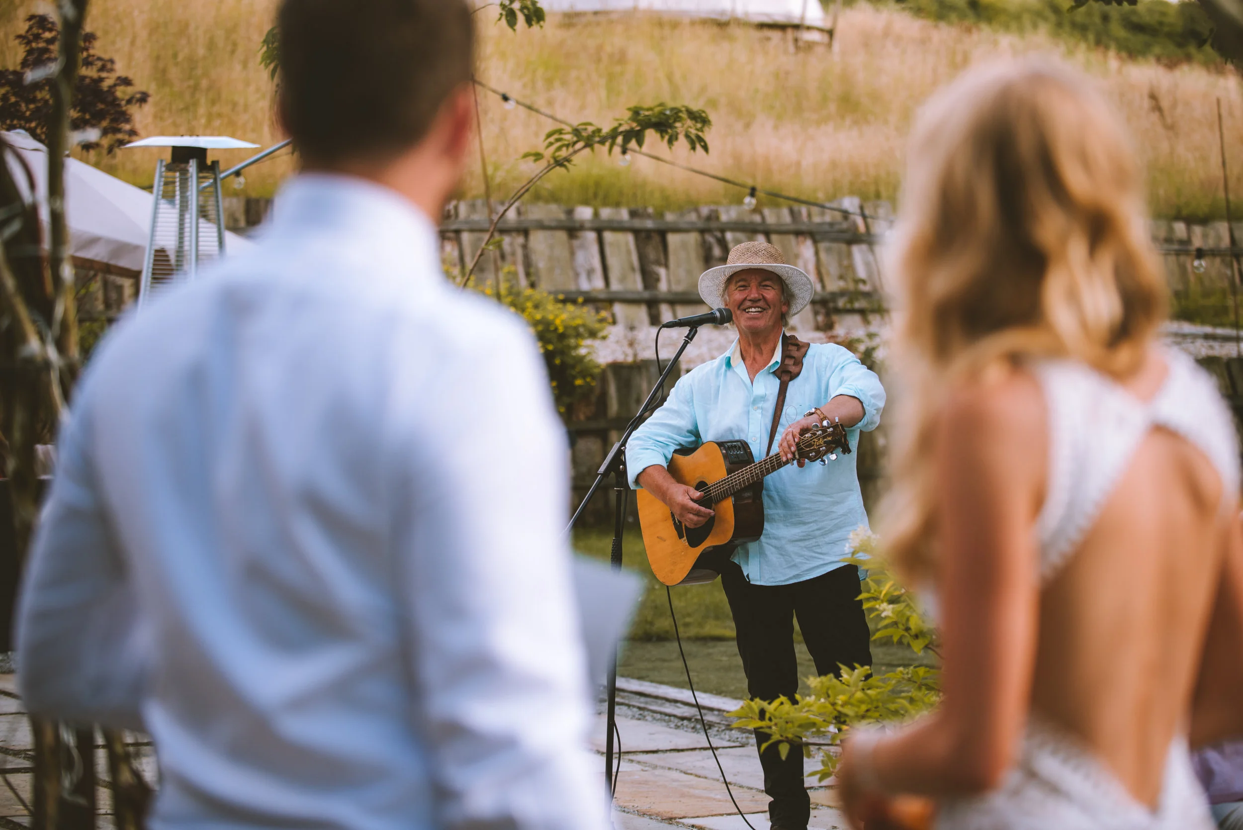 A man wearing a light blue shirt and a straw hat is playing an acoustic guitar and singing into a microphone at an outdoor gathering. Two people, a man and a woman, are seen from behind listening to him. The woman has long blonde hair and is wearing 
