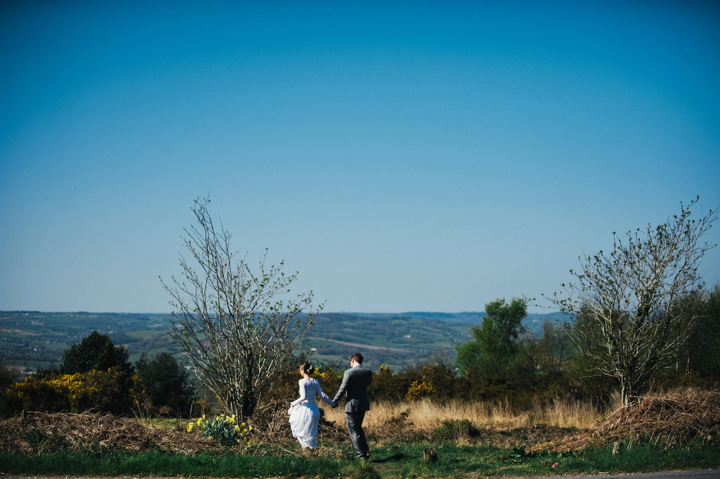 A couple walking hand in hand in a grassy, hilly landscape with trees and yellow flowers, under a clear blue sky.