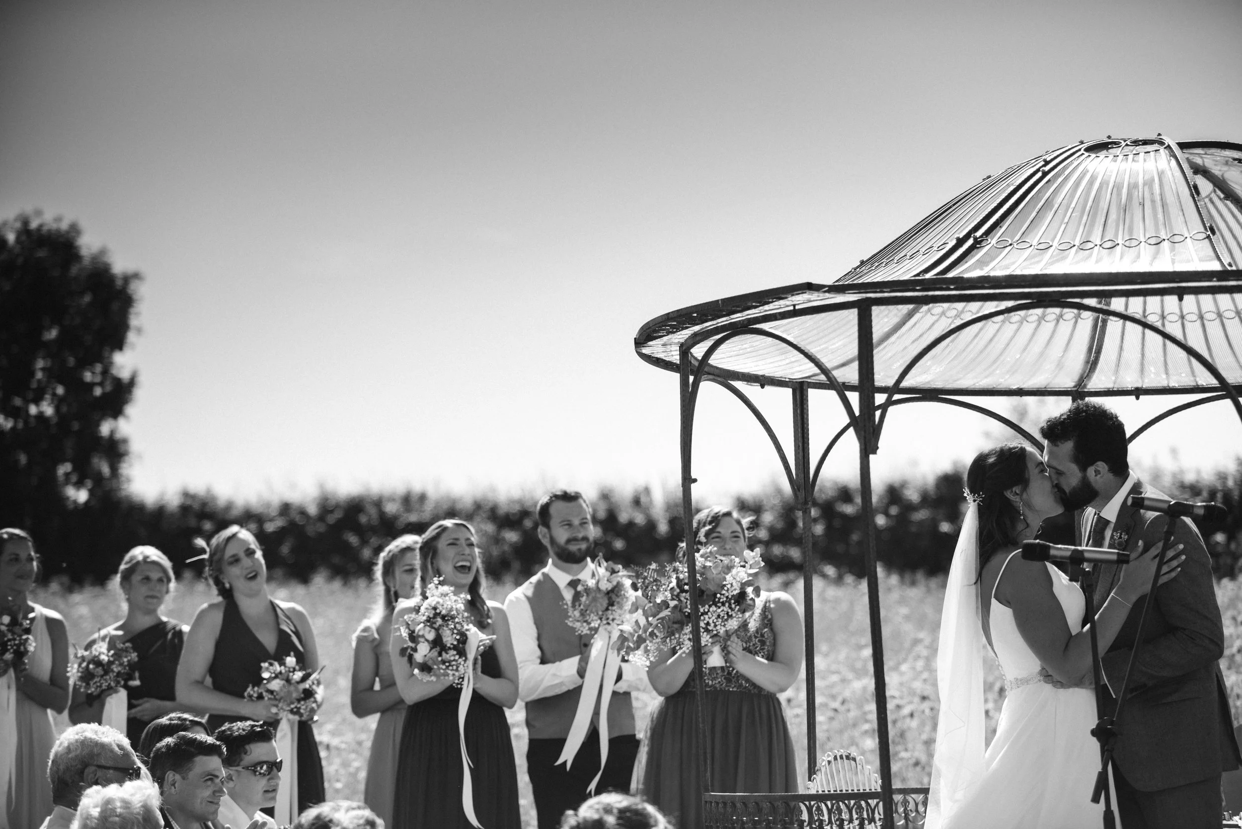 A black and white photo of a wedding ceremony outdoors with a group of people, including a bride and groom sharing a kiss under a decorative gazebo, surrounded by bridesmaids and guests watching and smiling.