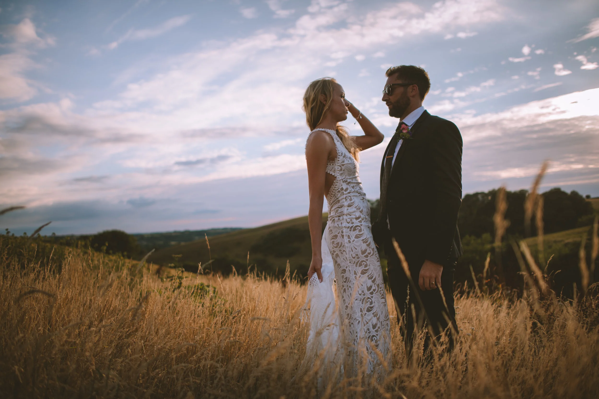 A bride and groom standing in a field of tall grass during sunset, facing each other closely, with a scenic landscape and cloudy sky in the background.