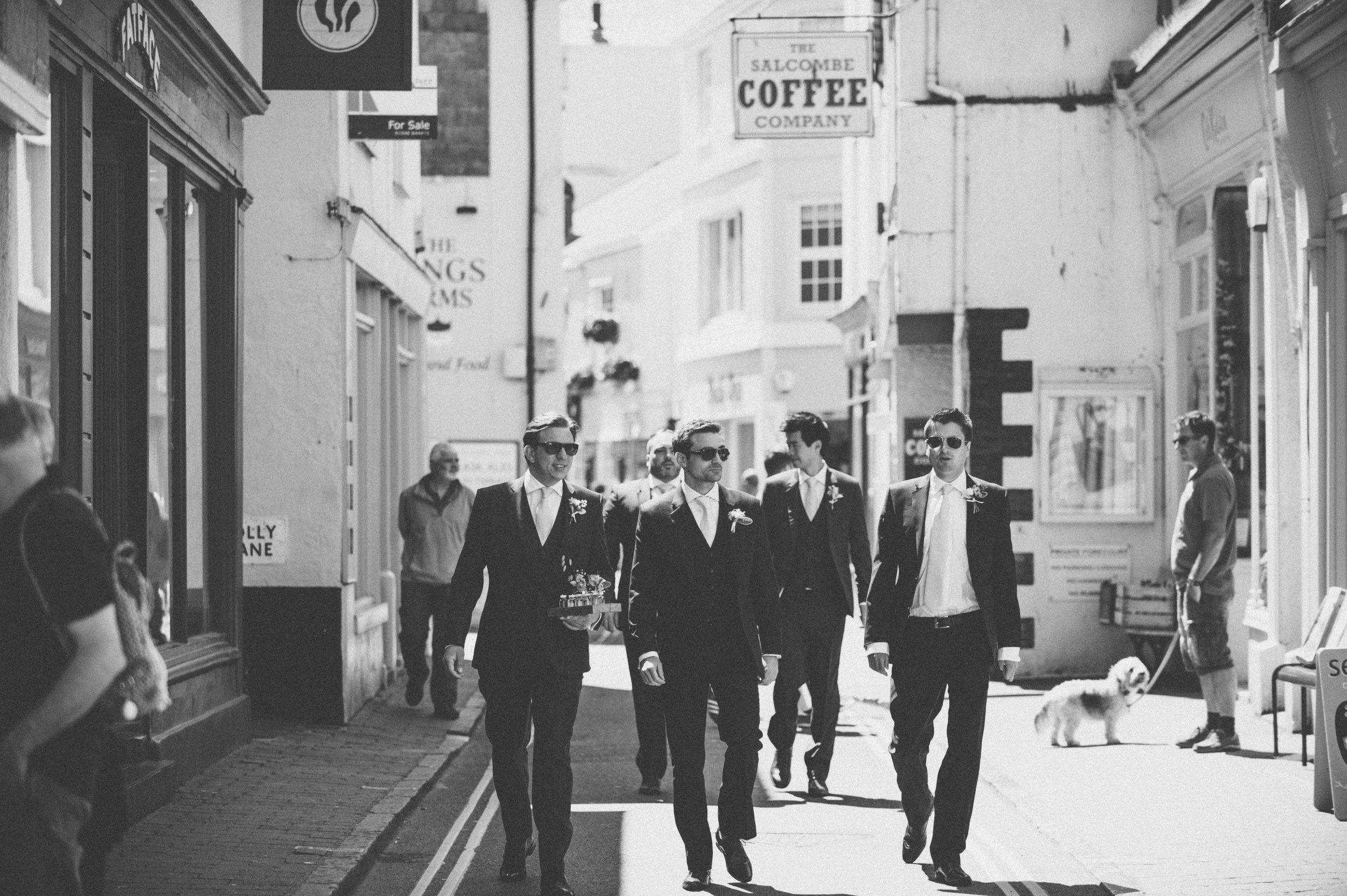 A group of men dressed in suits walking down a city street, some wearing sunglasses, with a dog in the background; black and white photo.