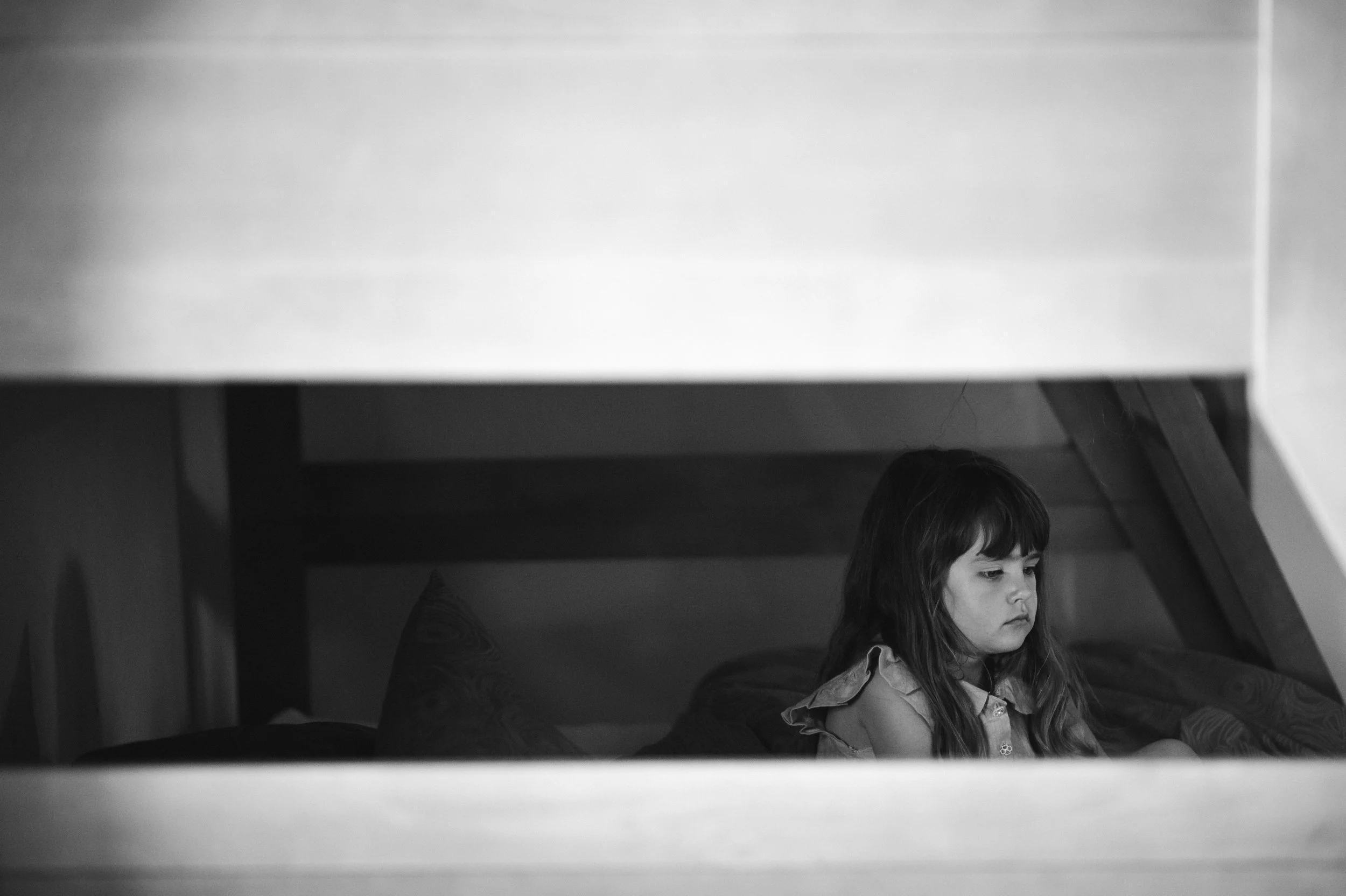 A young girl with long hair sitting on a bed, seen through a partially open door in black and white.