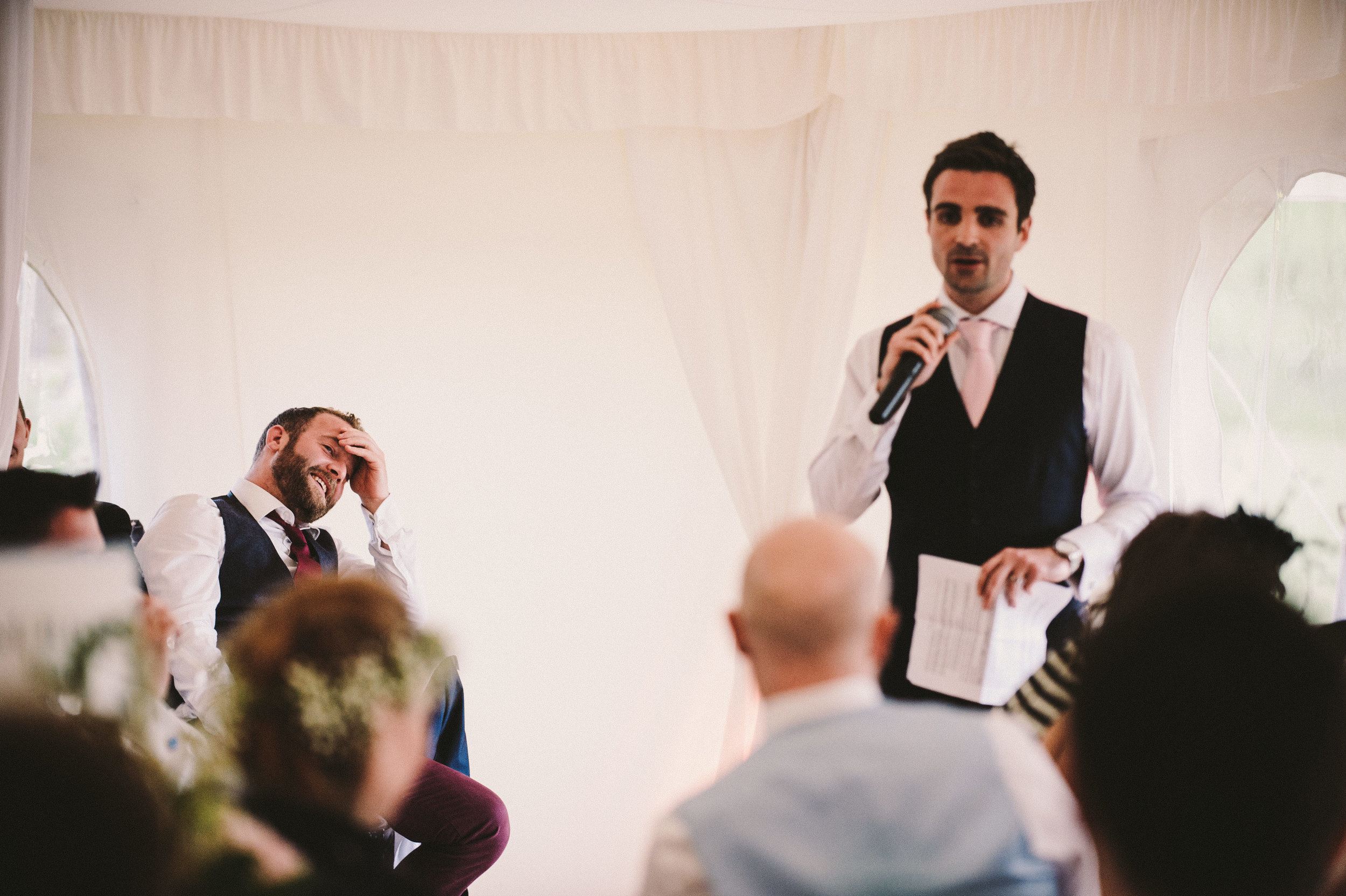 Man in suit giving speech at wedding reception, guest laughing with hand on forehead, others listening.