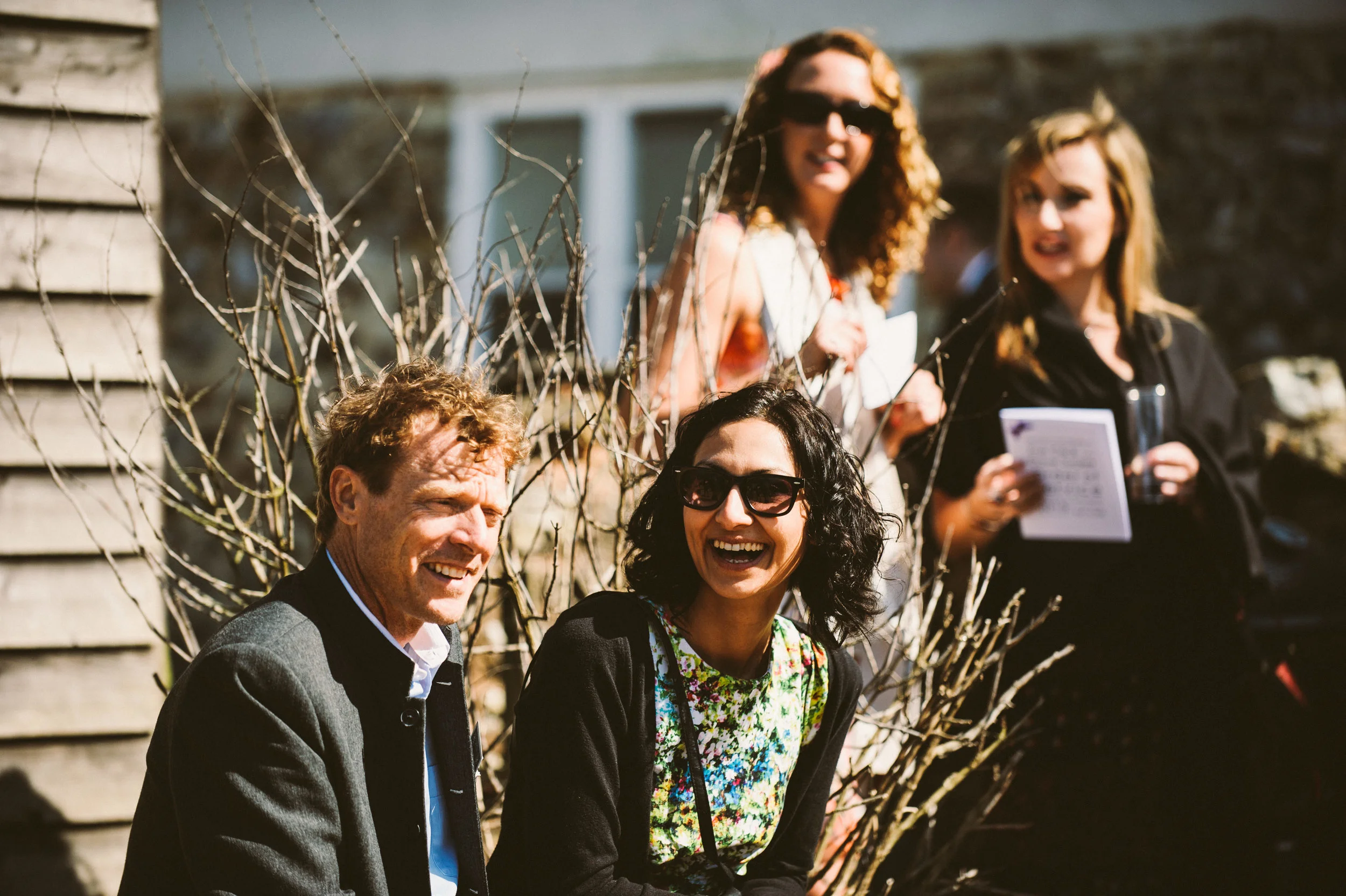 Four people outdoors, two sitting and two standing, in front of a leafless tree. The seated man has curly hair, wearing a dark blazer, smiling. The seated woman has black curly hair, wearing sunglasses and a floral dress, laughing. The standing woman