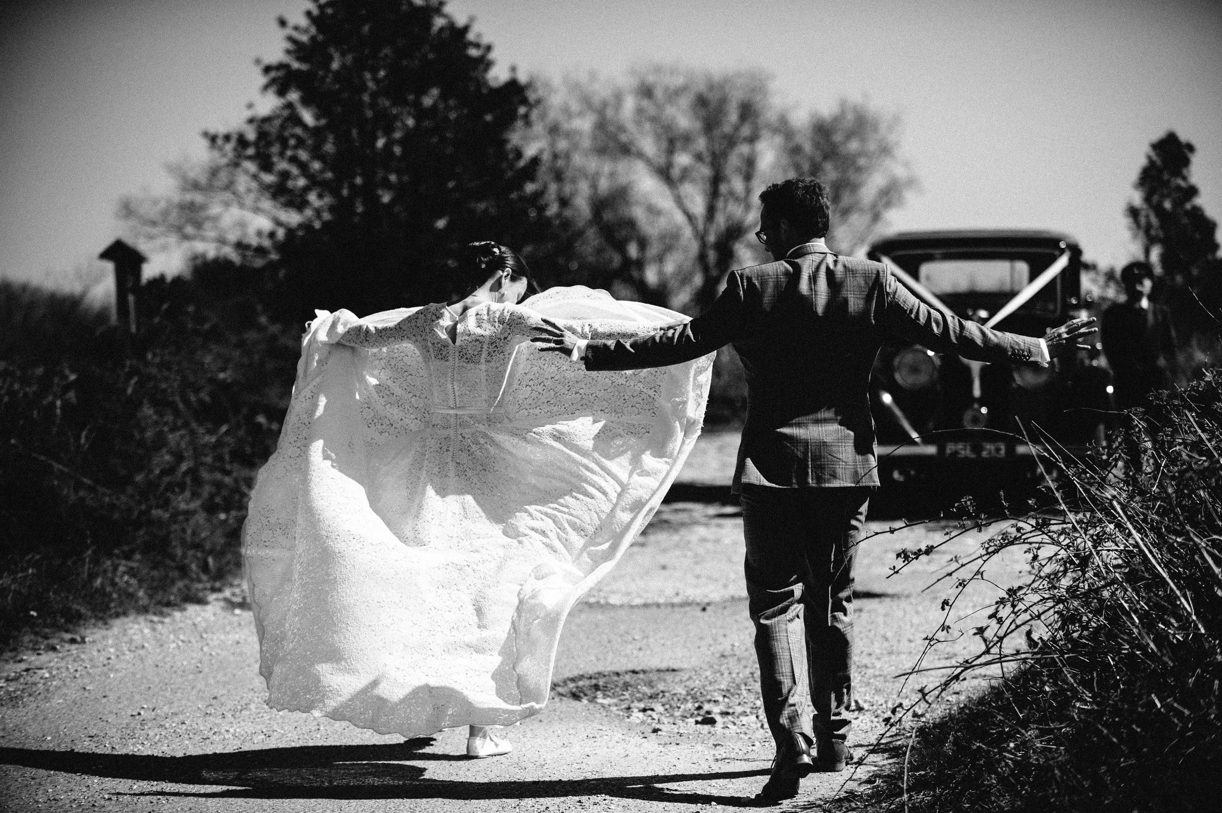 A black-and-white photo of a bride and groom walking hand in hand on a rural road. The bride is wearing a white gown and veil; the groom is in a plaid suit. An old car is visible in the background.