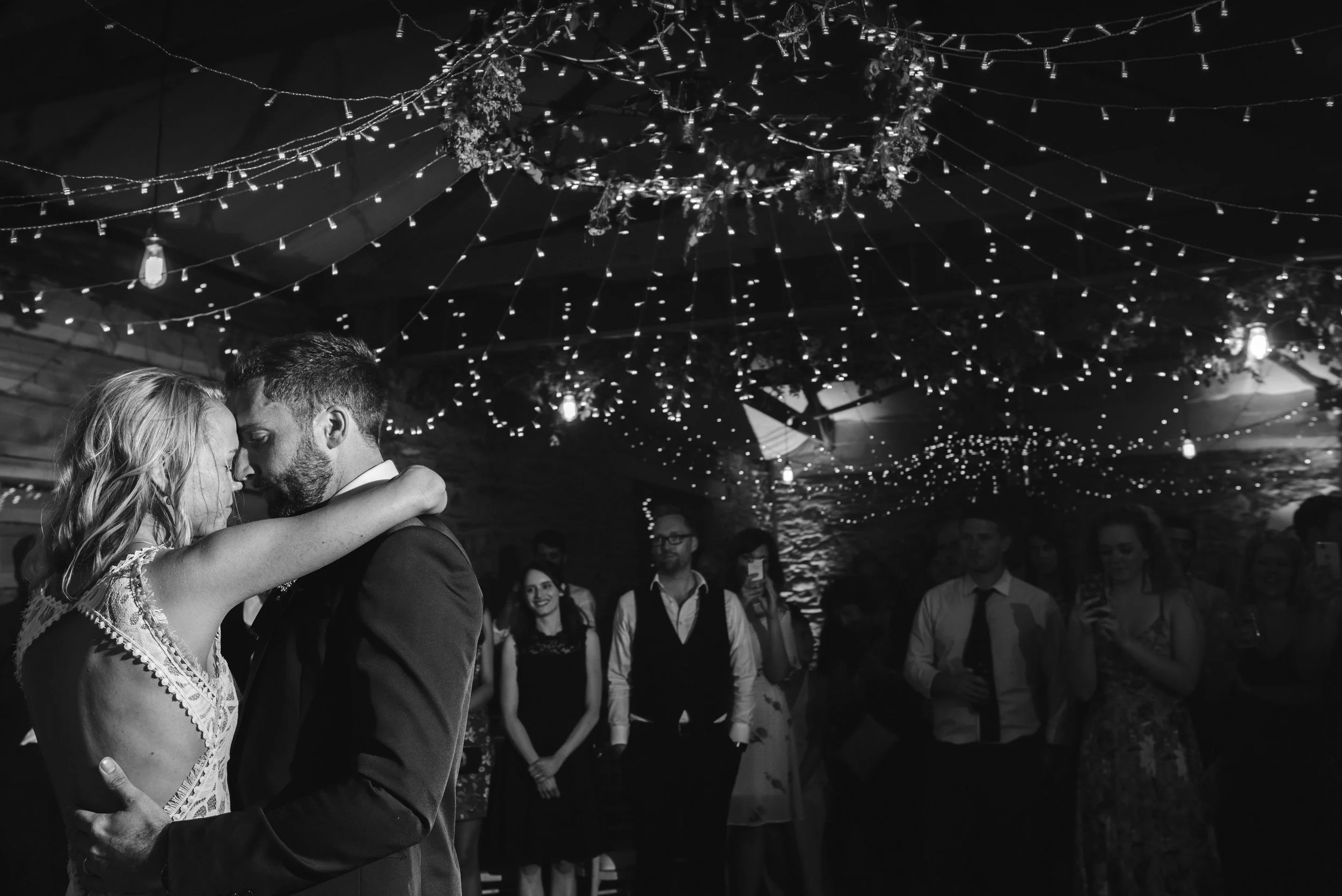 Black and white photo of a couple dancing closely at a wedding reception, under a ceiling decorated with string lights and hanging greenery, with guests watching in the background.