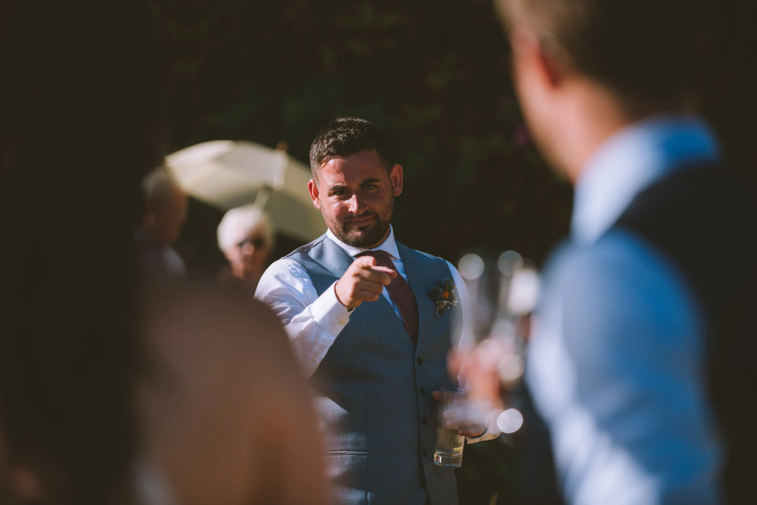 Man in a blue vest and white shirt pointing while holding a drink at an outdoor event, with other people and trees in the background.