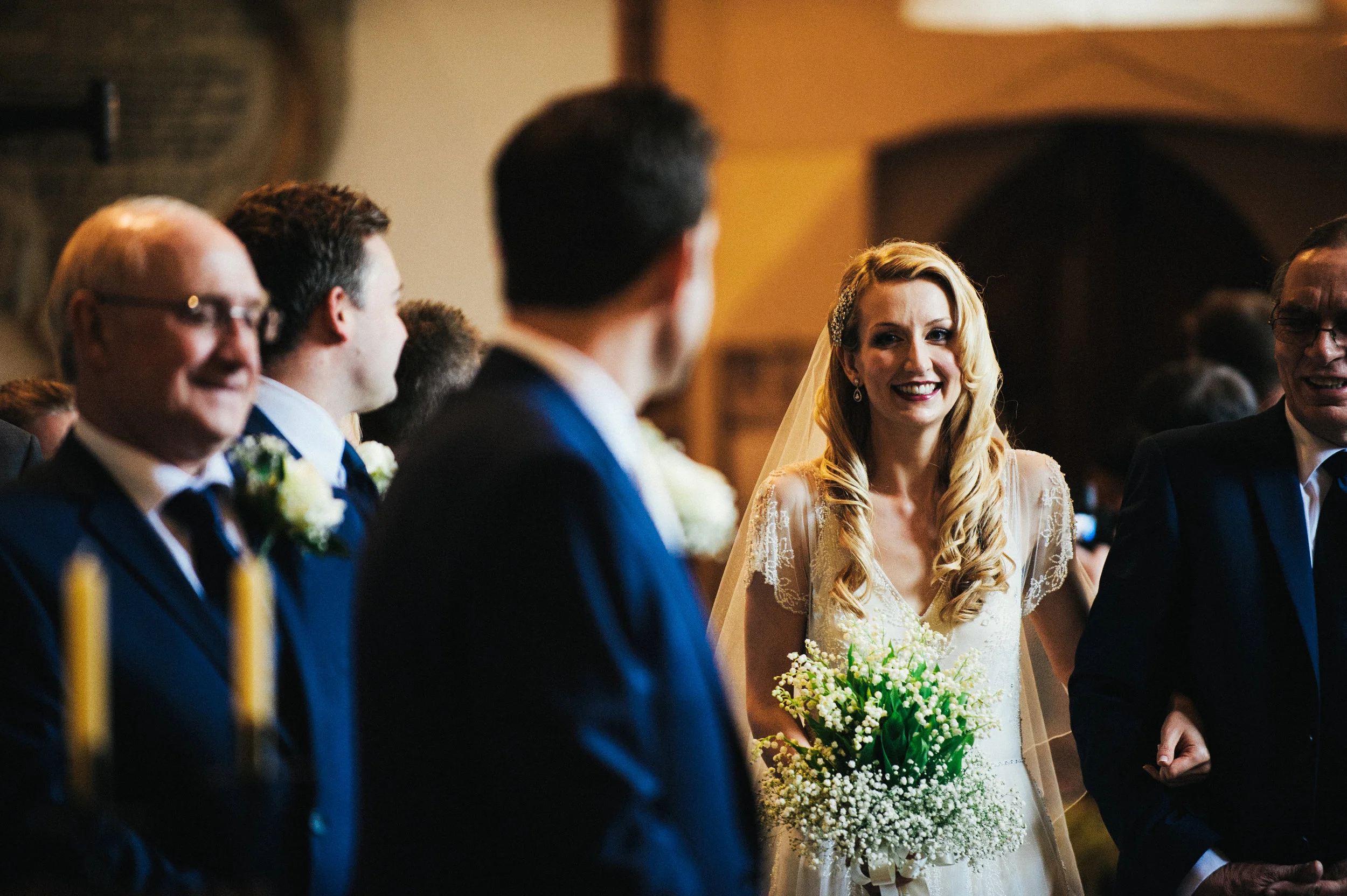 A bride with long blonde wavy hair in a wedding dress holding a bouquet of white flowers, smiling at a wedding ceremony surrounded by men in suits.