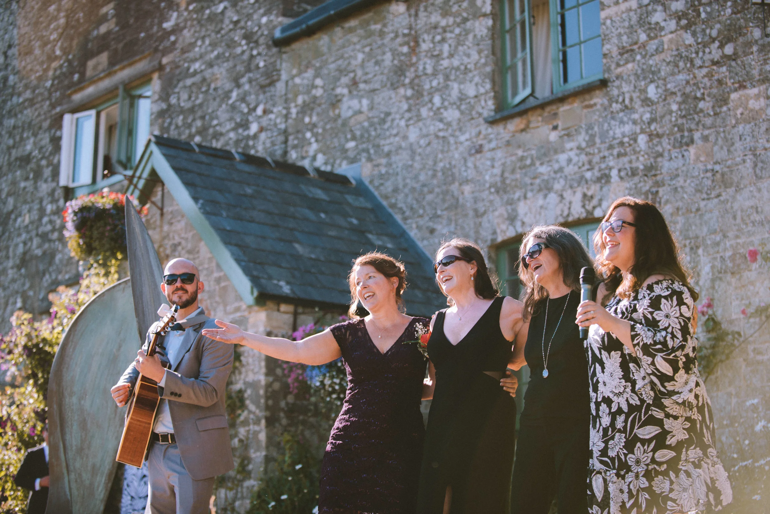 A group of five people standing outdoors in front of a stone building, smiling and singing together. One man is playing a guitar, and a woman on the right is holding a microphone. They appear to be enjoying a musical performance or celebration.