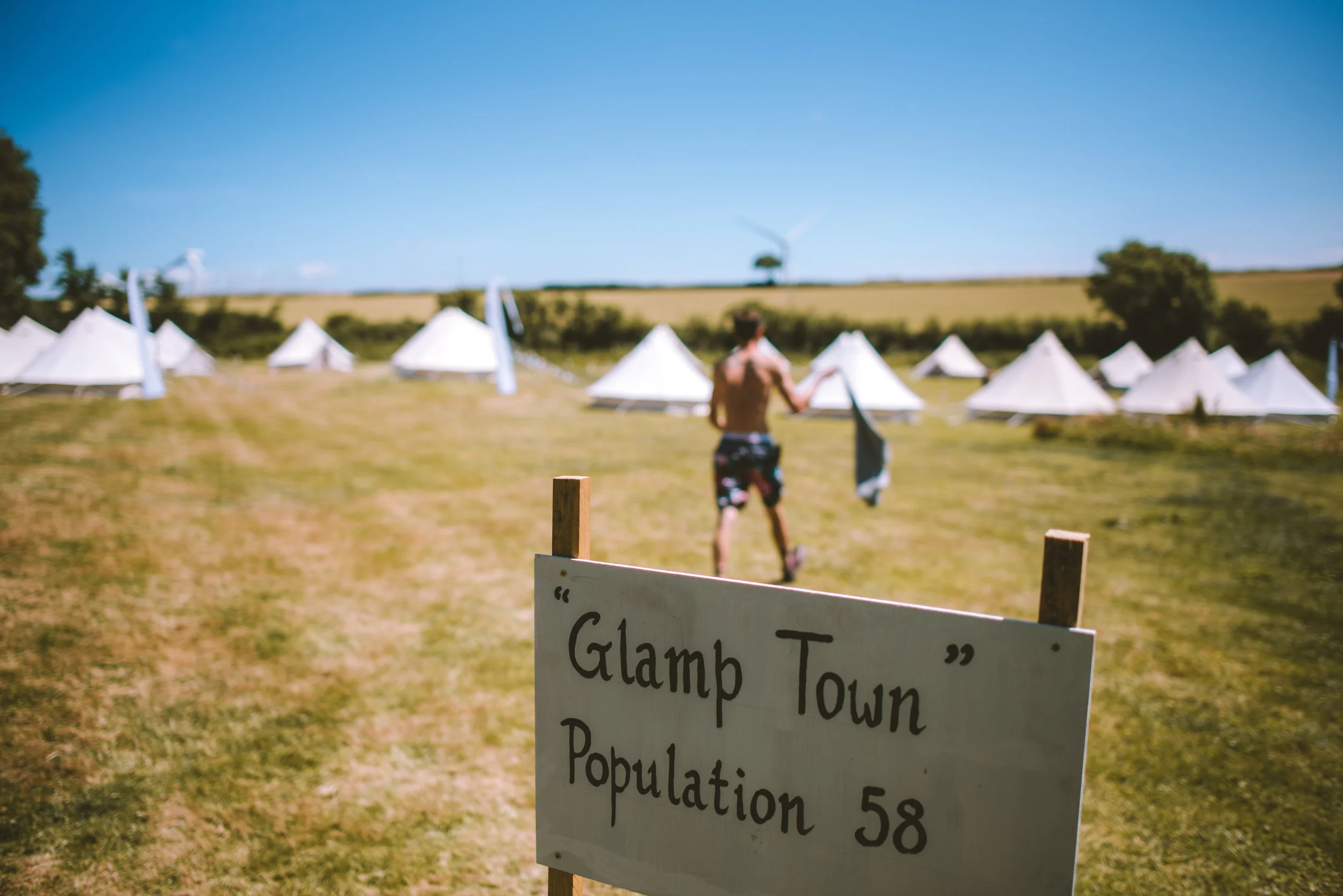 A man in swim trunks walking through a field with white tents in the background, a sign in the foreground reads 'Glamp Town', Population 58.
