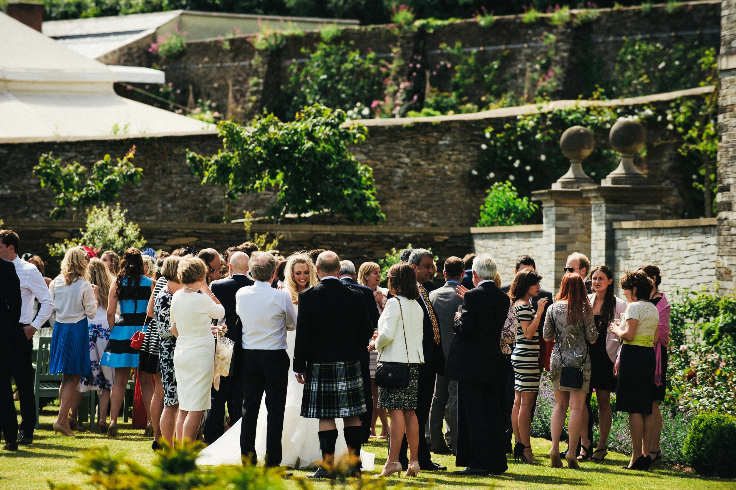 Group of people dressed in formal attire, gathered outdoors on a lawn, at a social event or celebration, with stone walls and greenery in the background.