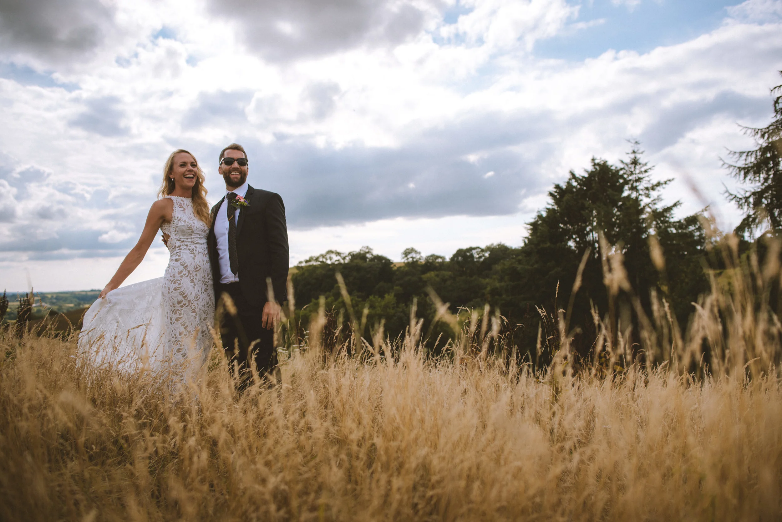 A happy couple in wedding attire standing in a field of tall grass with trees and a partly cloudy sky in the background.