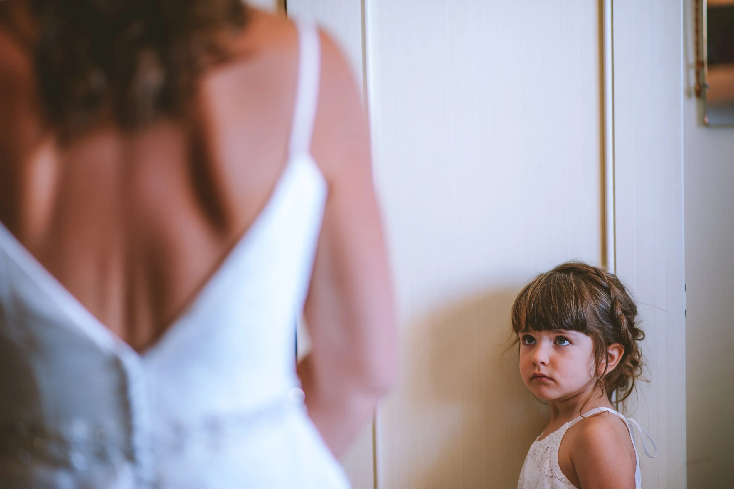 A young girl with brown hair and bangs looking at a woman, possibly her mother, who is partially visible in the foreground. The girl wears a white dress with lace and tied shoulder straps, standing against a neutral-colored wall.