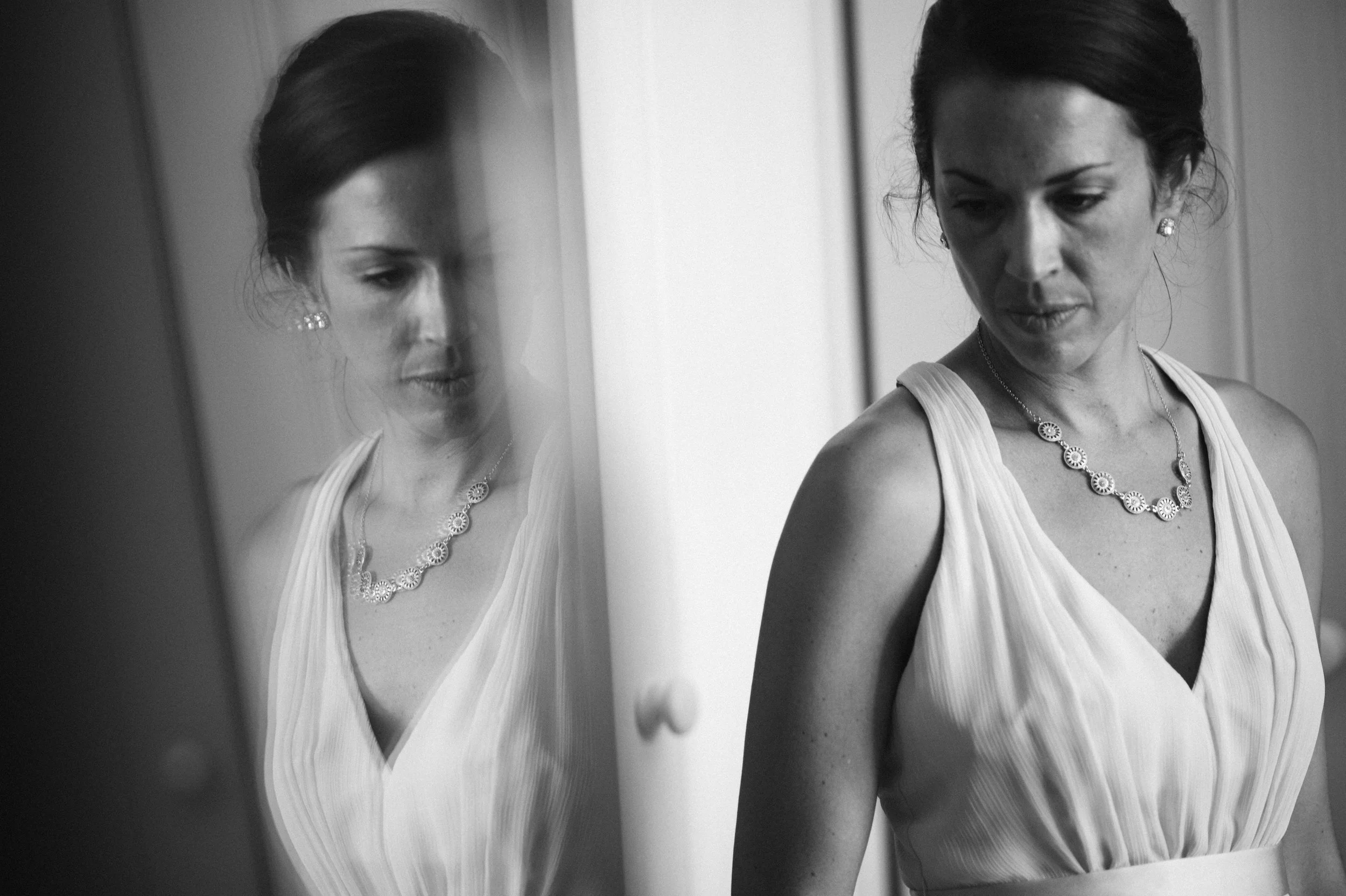 A woman in a sleeveless, V-neck dress looking down, with her reflection visible in a mirror.