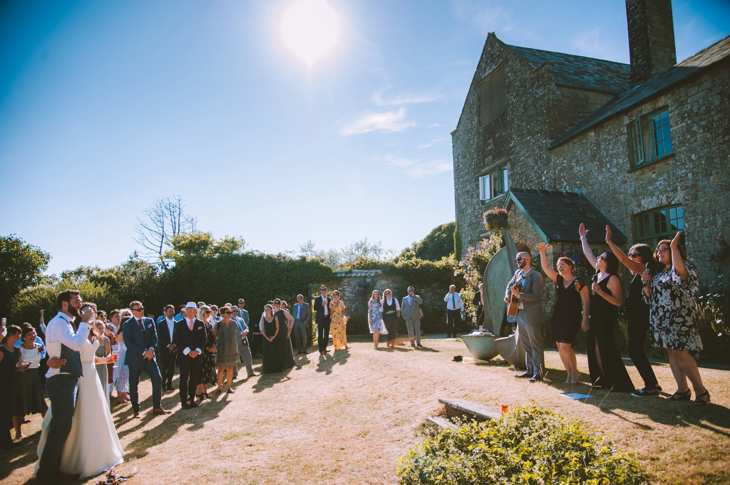 People gathered outdoors at a wedding, with a bride and groom in the foreground and a band performing on the right, with an old stone building and clear blue sky in the background.