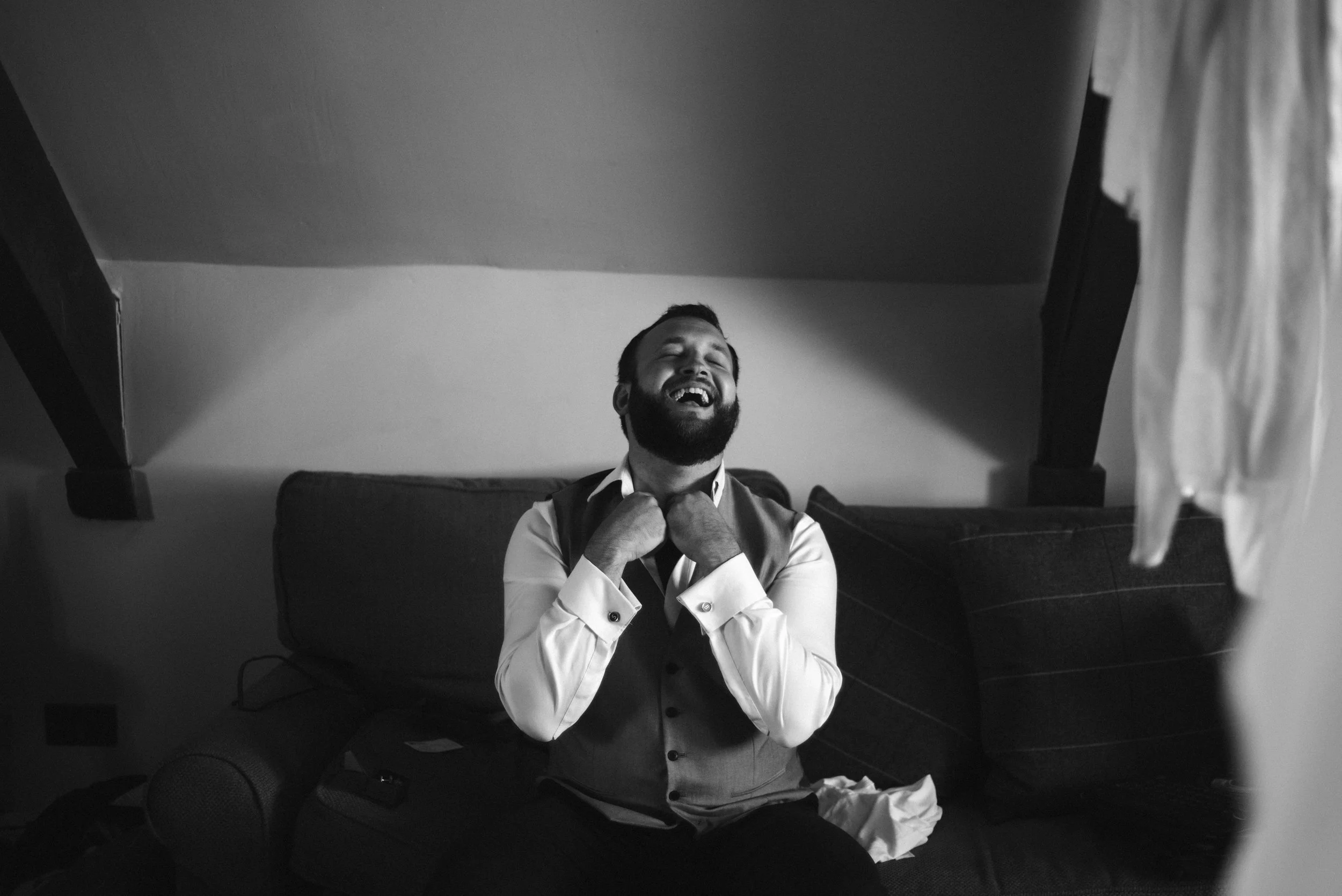 A man with a beard laughing and adjusting his tie while sitting on a couch in a room with sloped ceiling, black and white photo.