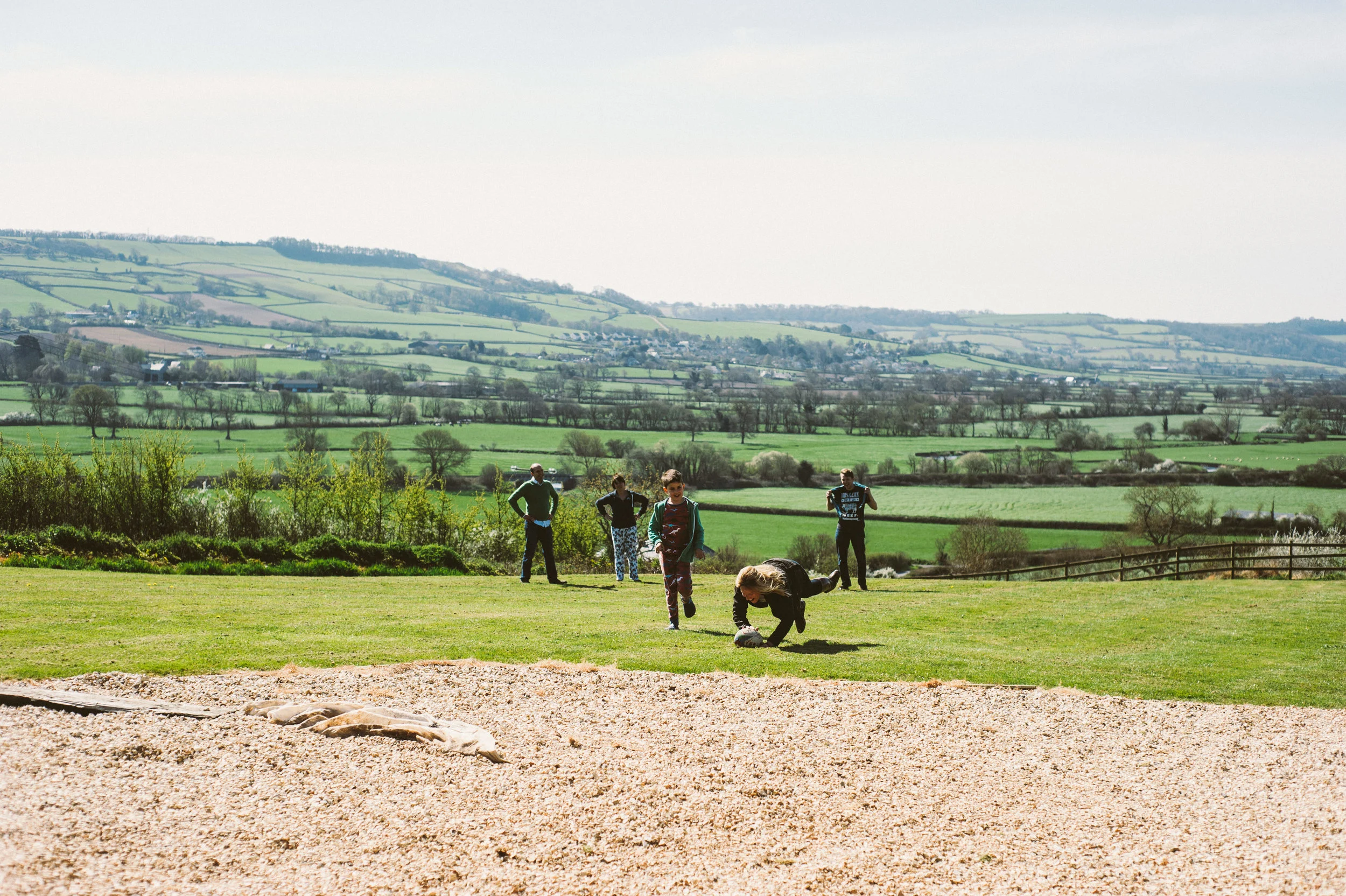 A group of people, including a girl, are playing rugby outdoors on a grassy field with a scenic countryside background of rolling hills and trees.