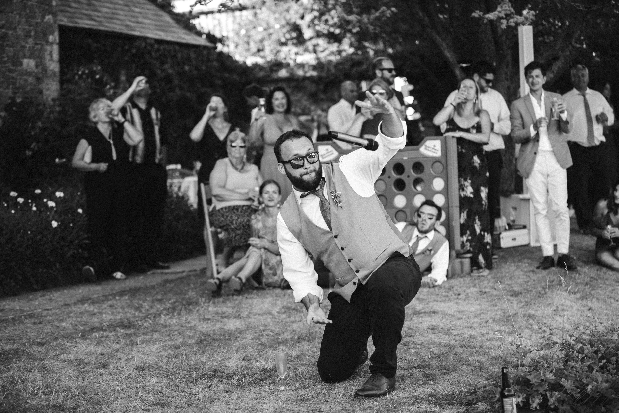 A man with sunglasses and a vest is dancing at an outdoor celebration, surrounded by a group of casually dressed people watching and enjoying the event.