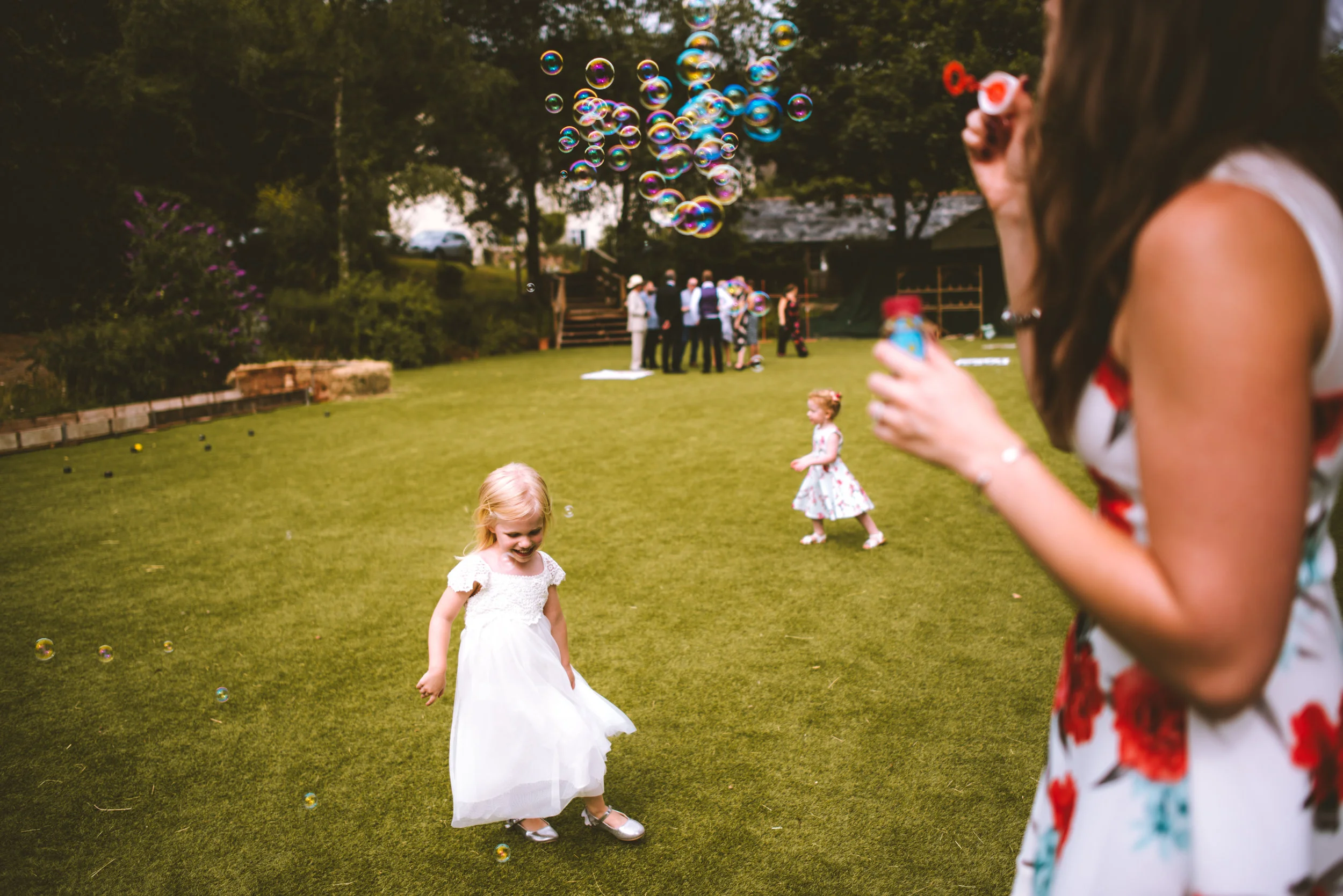 A young girl in a white dress playing with bubbles on a green lawn during a gathering, with a woman blowing bubbles on the right side of the image and a group of people in the background.