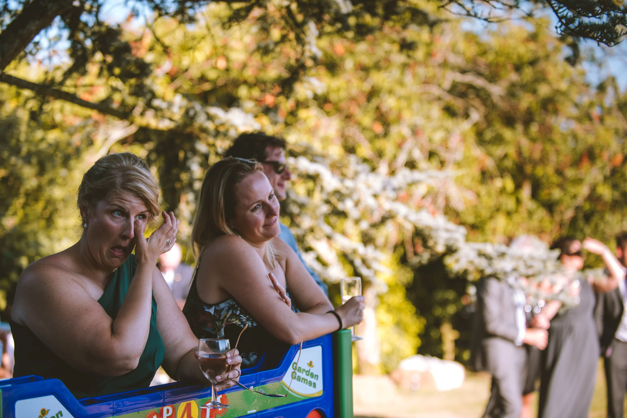 Group of people at an outdoor event, some standing and others sitting, with trees and flowers in the background, enjoying drinks and socializing.