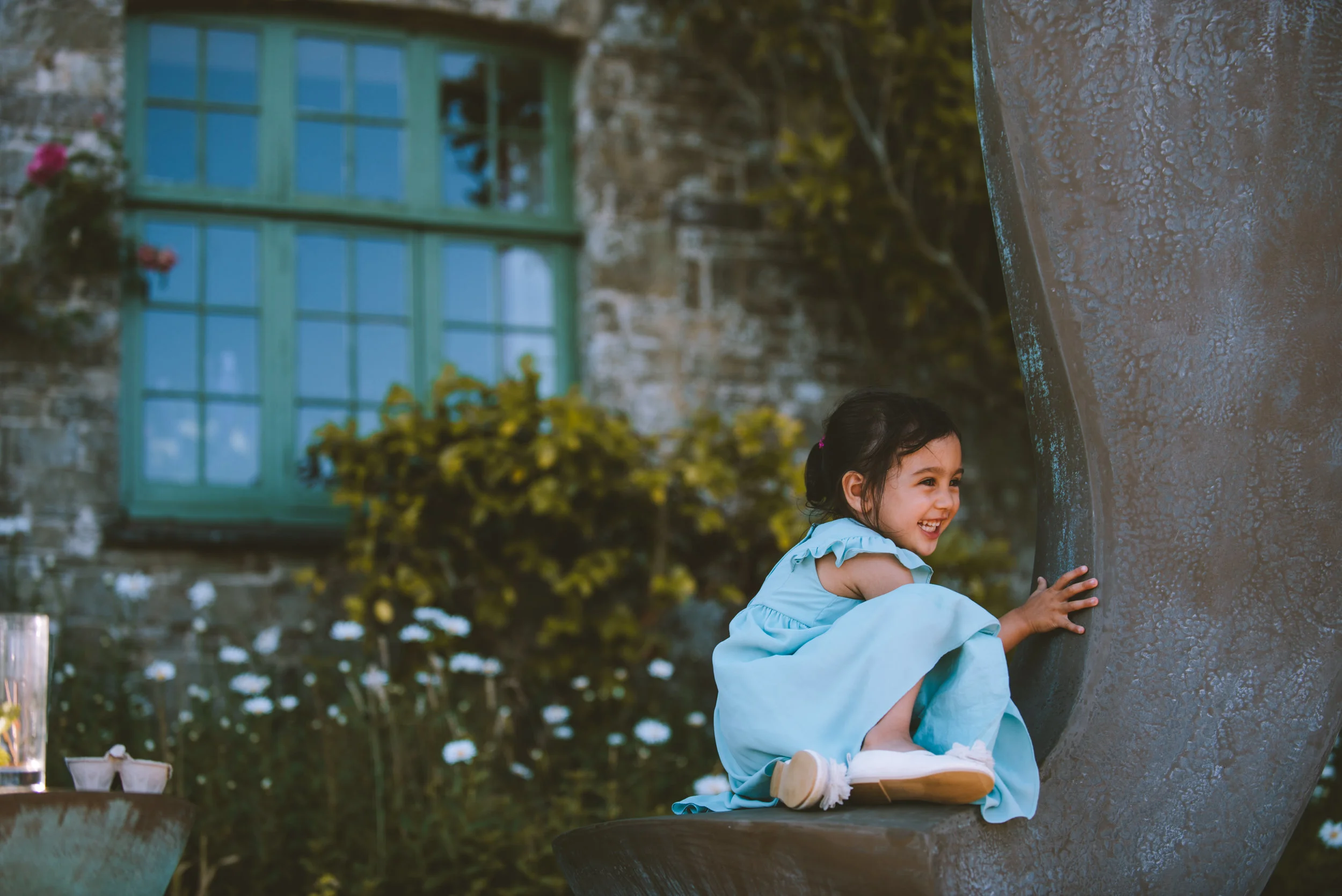 A young girl in a light blue dress sitting on a large curved outdoor sculpture, smiling and playing, with a garden and a green windowed building in the background.