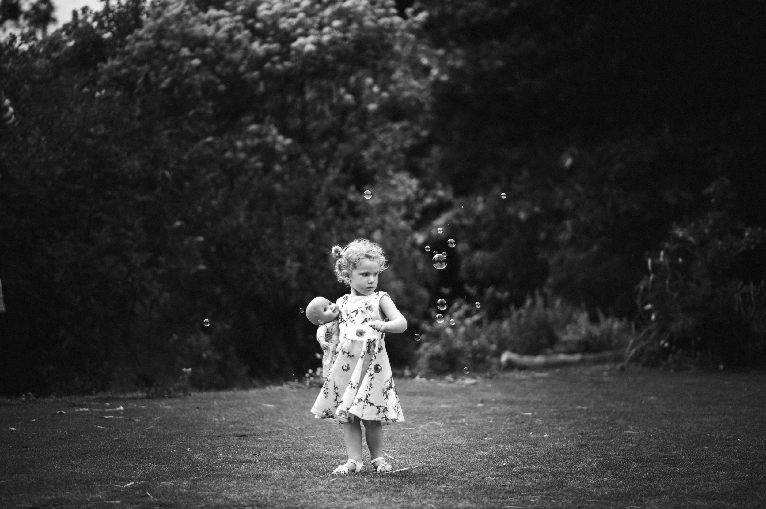 A young girl with curly hair in a floral dress stands outdoors on a grassy area, holding a doll, with bubbles floating around her and trees in the background.