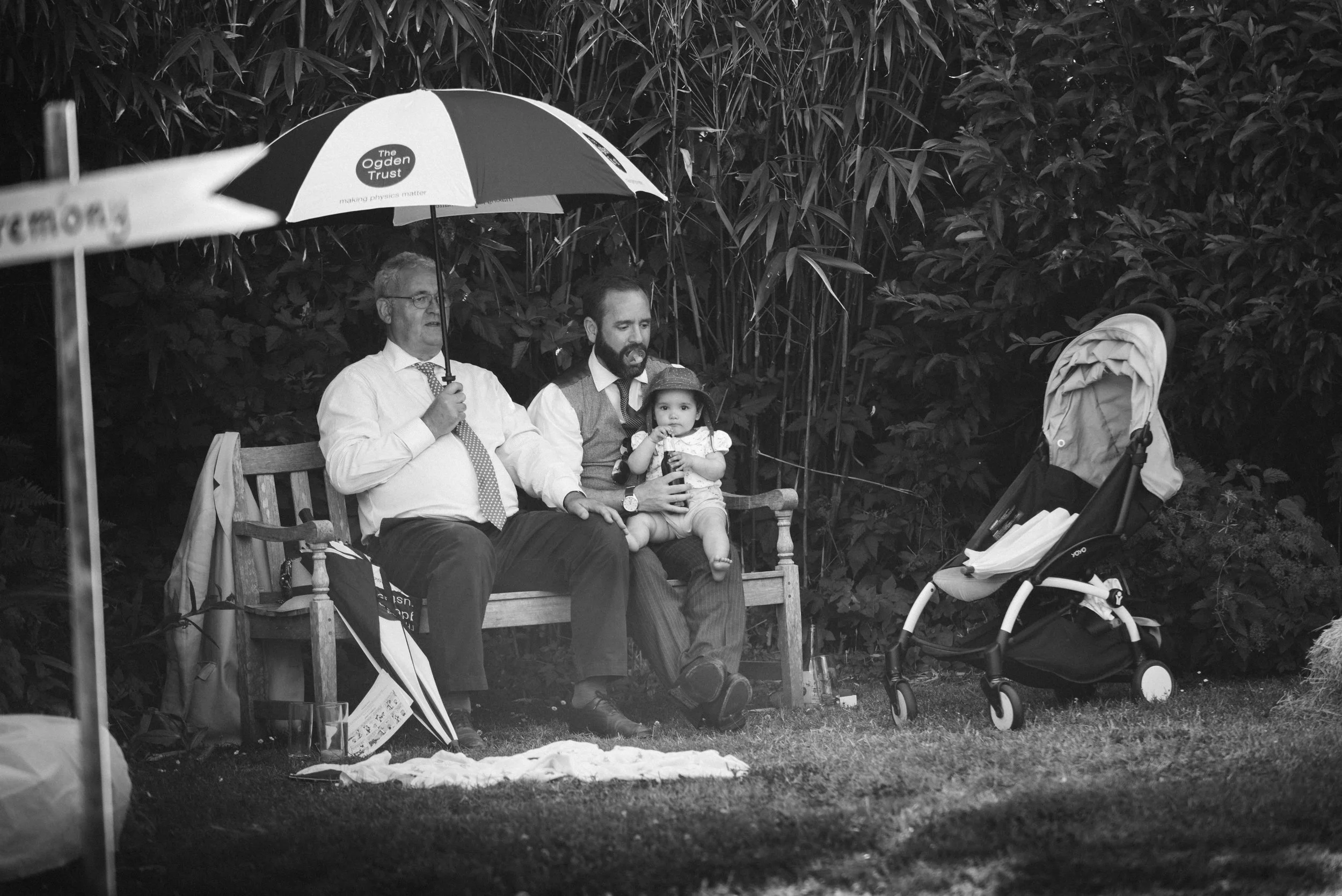 Three people sitting on a park bench, with an umbrella, a baby, and a stroller nearby. A man holds the umbrella, an older man and a young man sit together, and the baby is sitting on the young man's lap, all in front of dense foliage.