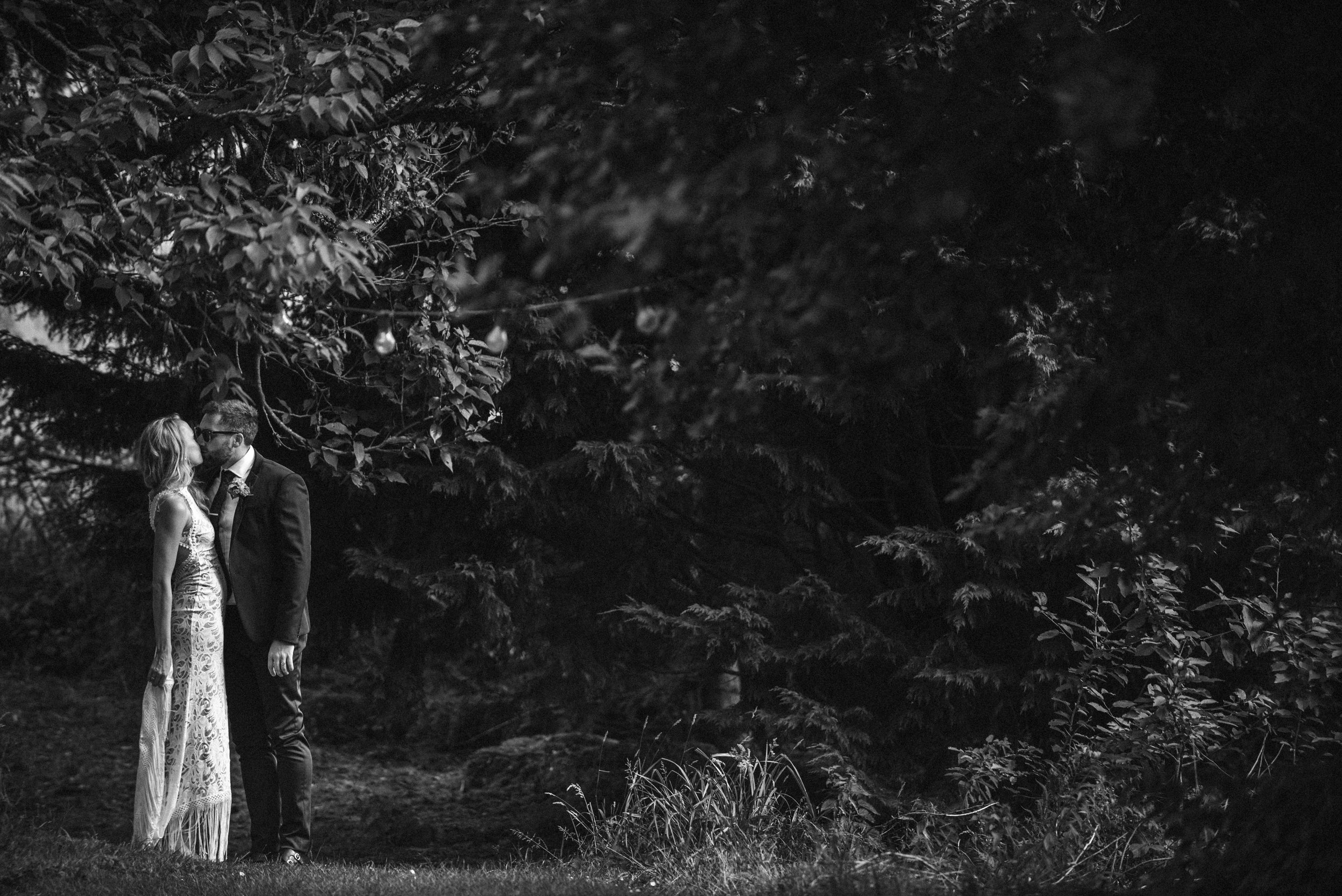 A black and white photograph of a couple kissing outdoors at night, surrounded by trees and foliage.