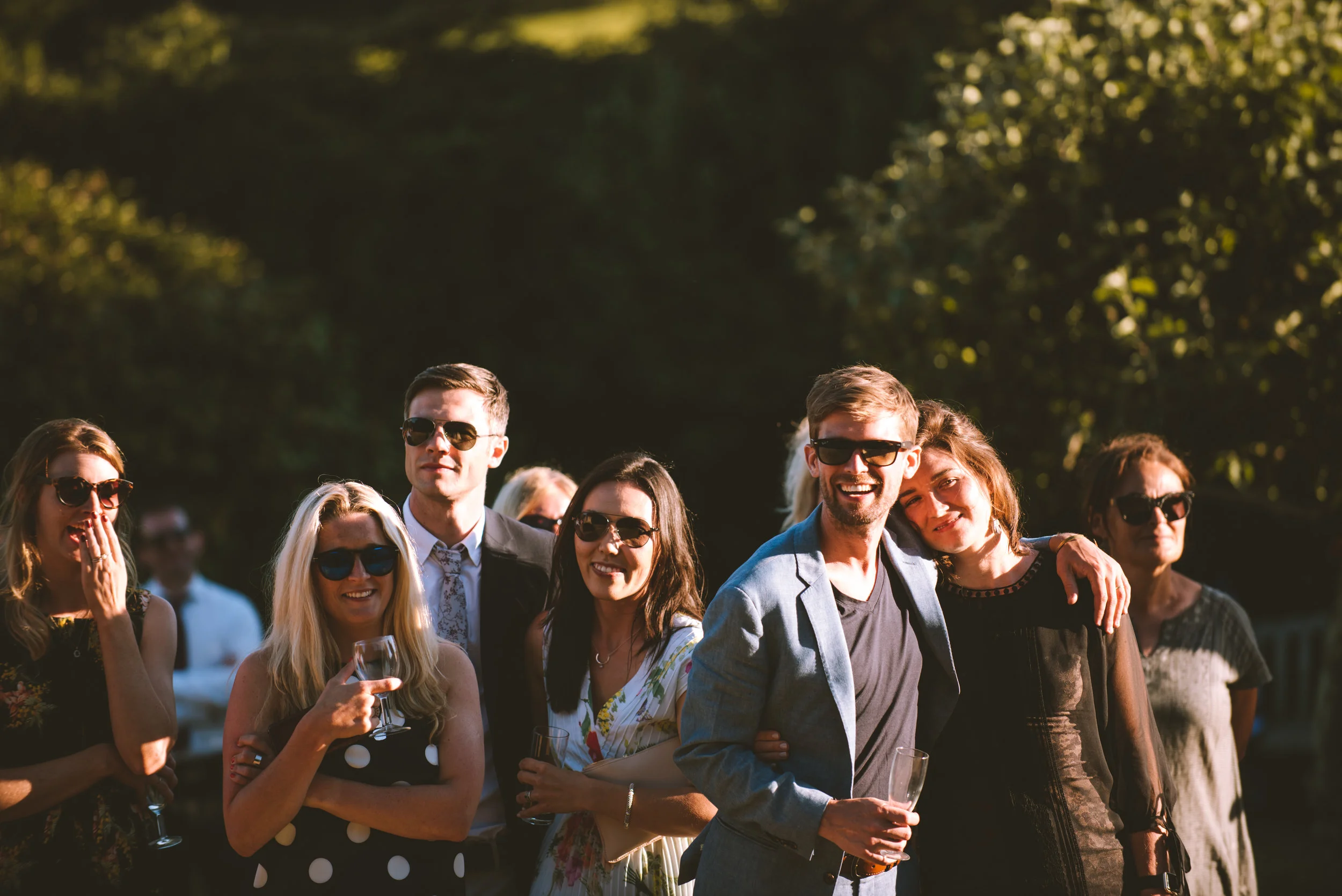 Group of well-dressed people at an outdoor event, wearing sunglasses and holding glasses of wine or champagne, smiling and enjoying the sunny day.