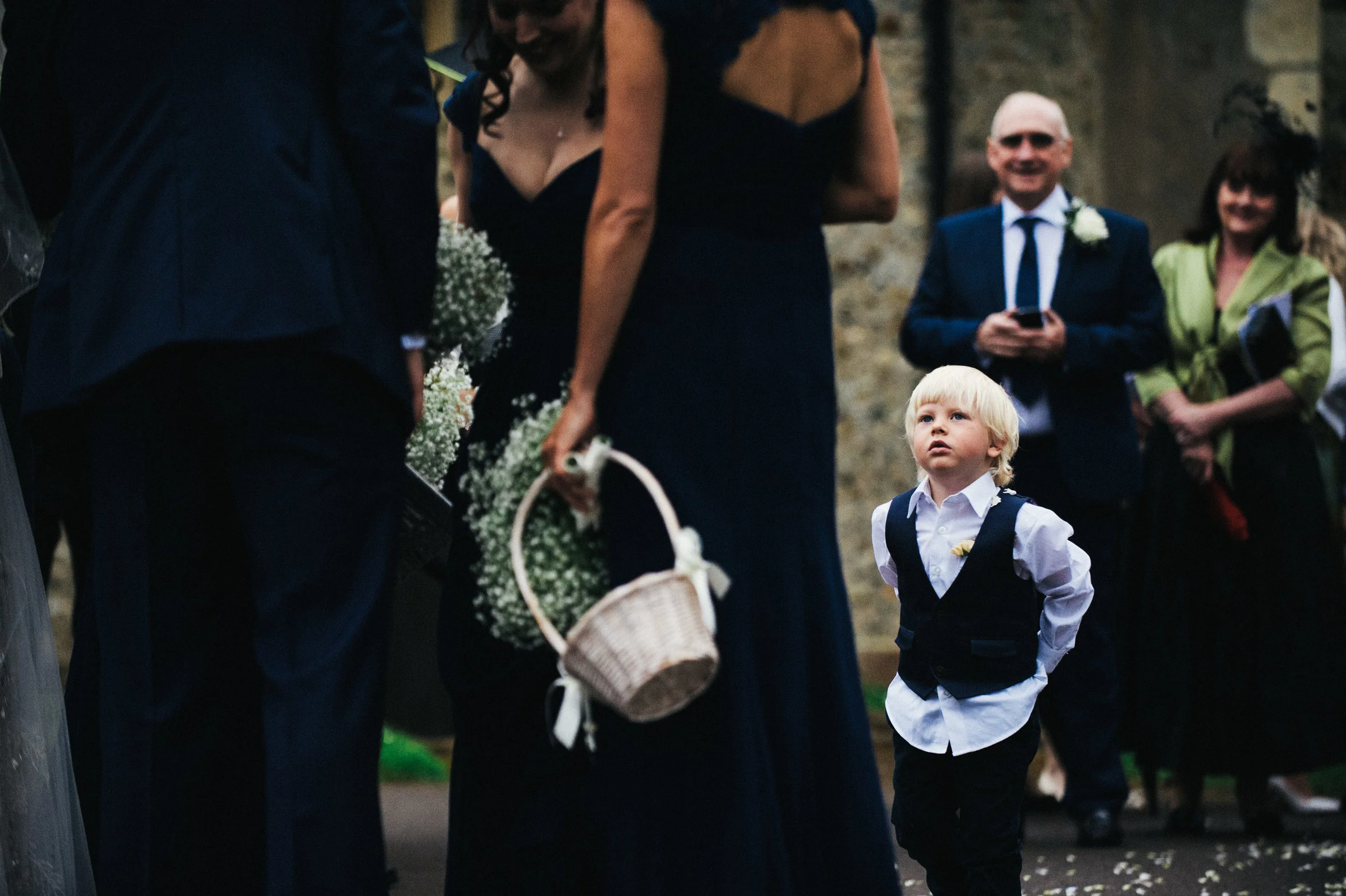A young boy with blonde hair, dressed in formal attire, standing among adults at a wedding or formal event, looking upward.