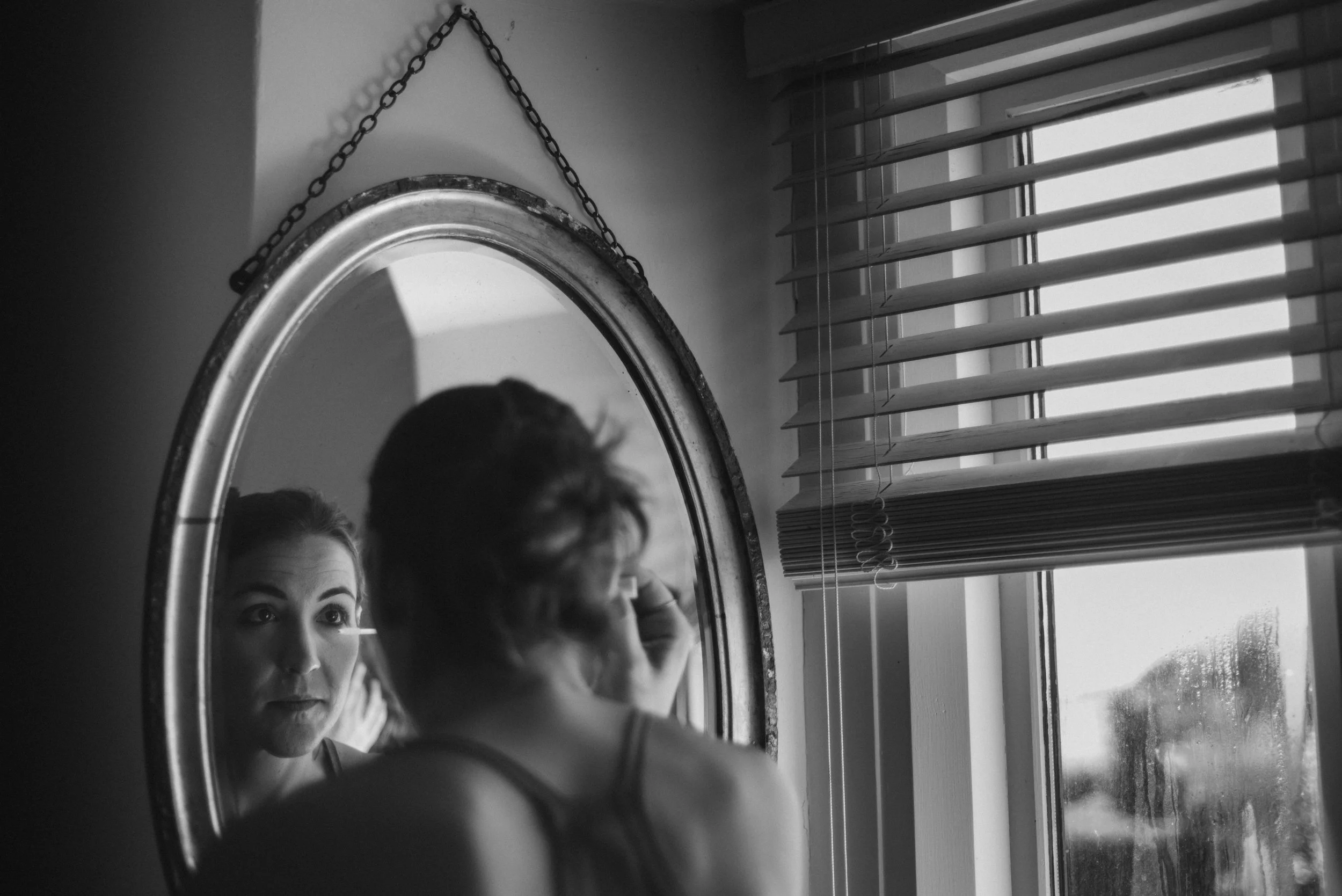 A woman looking at herself in a round mirror hanging on the wall, with window blinds partially open beside her.