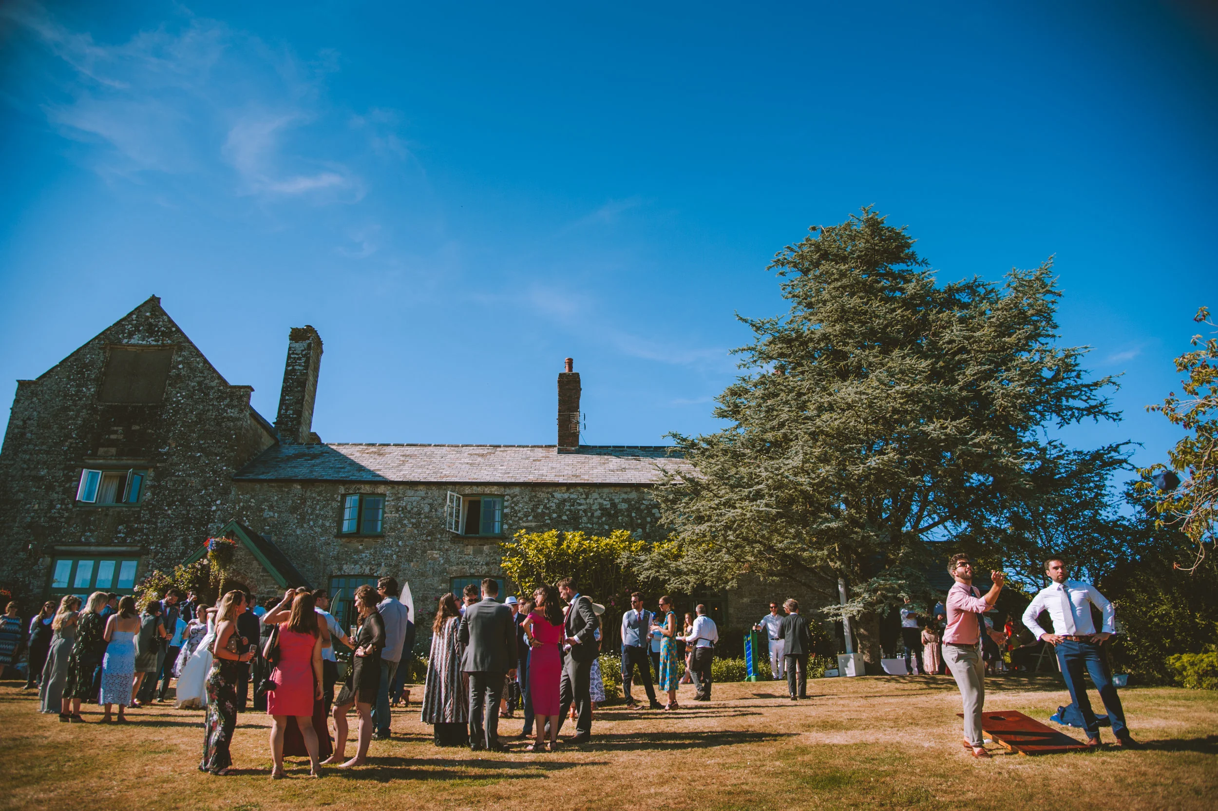 People gathered outside a stone building with a large tree, under a bright blue sky on a sunny day.