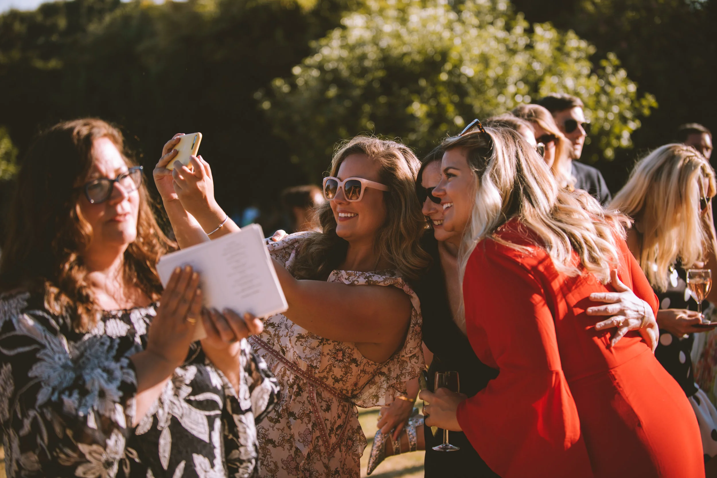 A group of women are taking a selfie outdoors during daylight. They are smiling and holding glasses of wine. The background shows trees and other people at a social event.