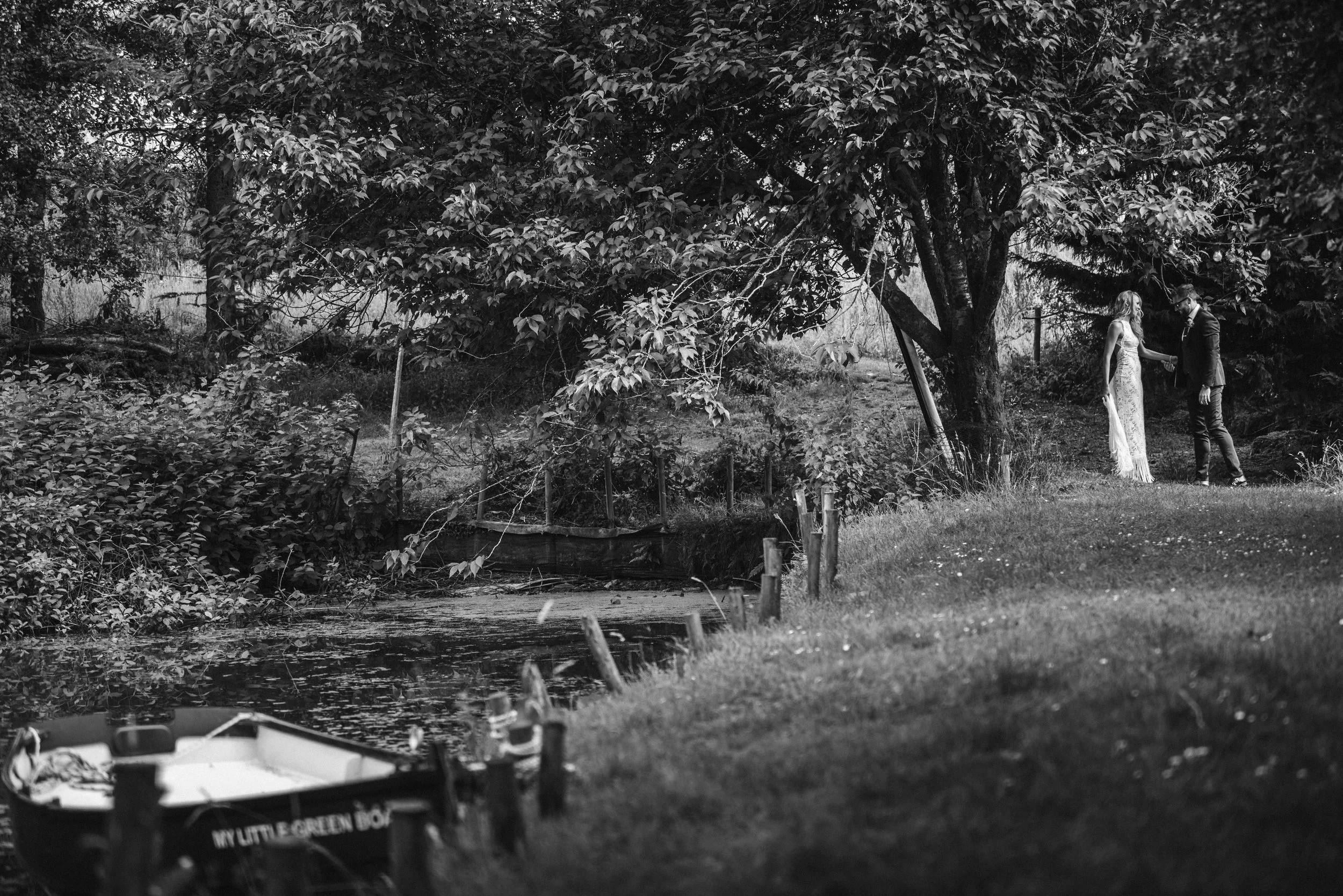 A black and white photo of a couple in formal attire holding hands near a large tree by a small pond in a park or garden setting.