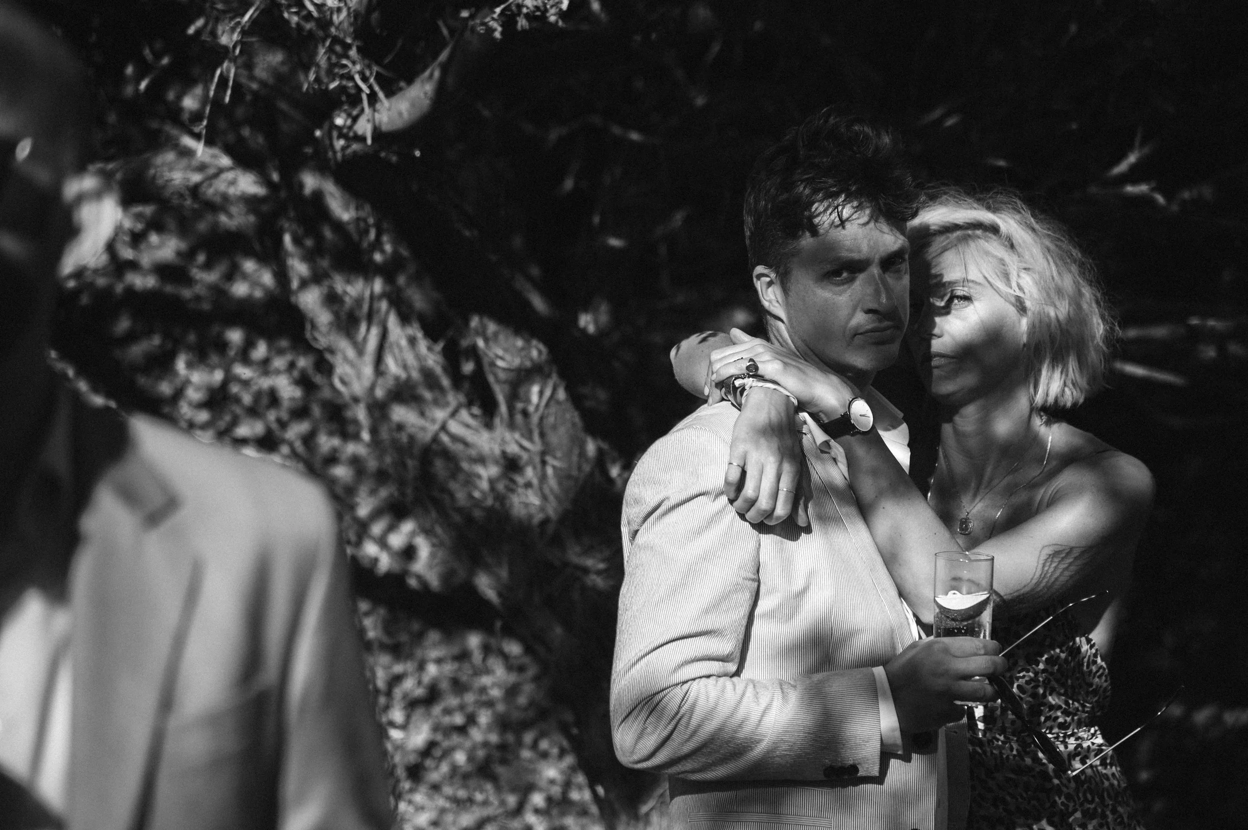A black-and-white photo of a man in a light-colored suit holding a drink, with a woman with short, curly blonde hair and a tattoo on her arm embracing him. The woman is smiling and looking at the camera, while the man is looking at the camera with a 