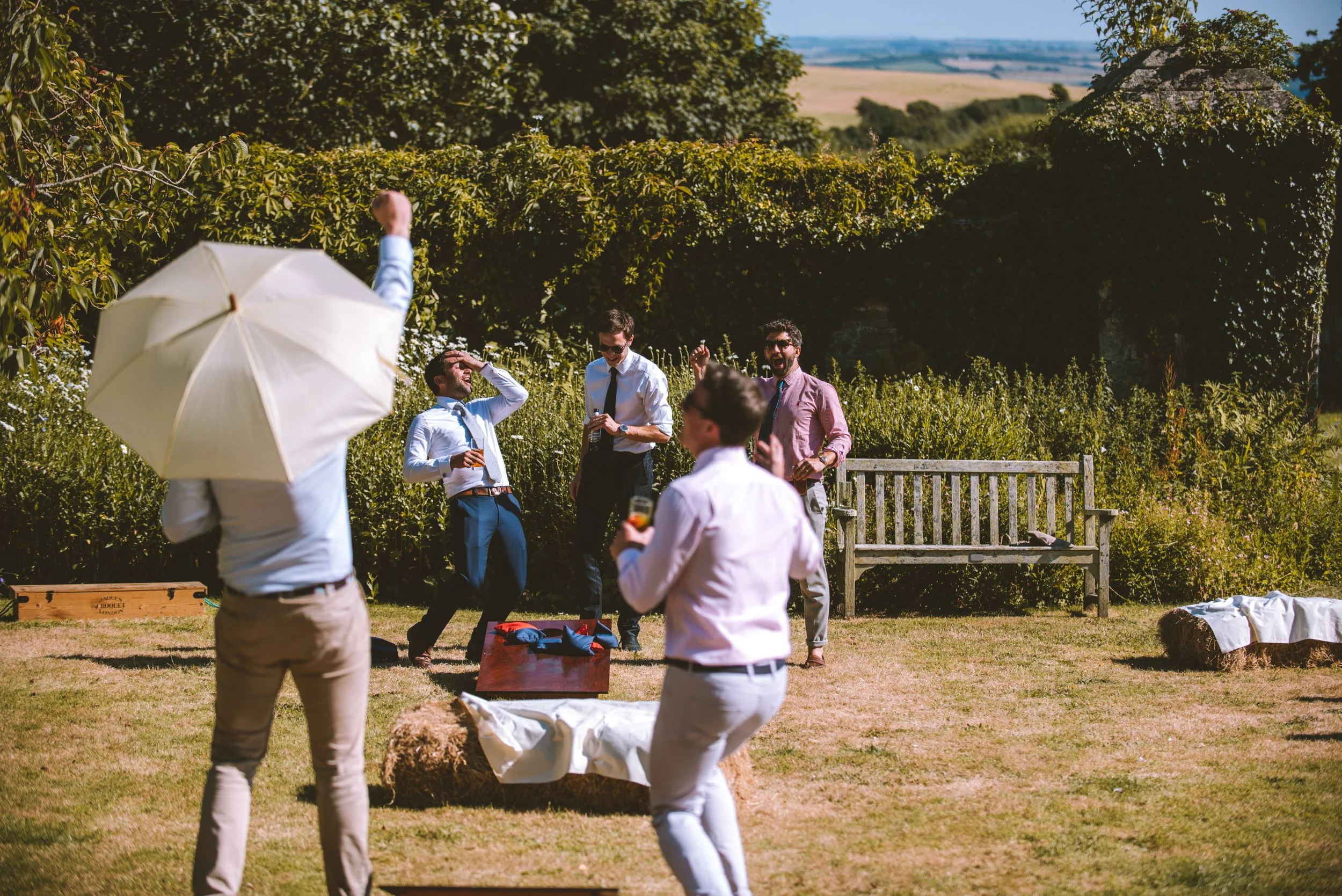 Group of men enjoying drinks and laughs outdoors on a sunny day, surrounded by trees and a grassy field, with a bench and hay bale visible.