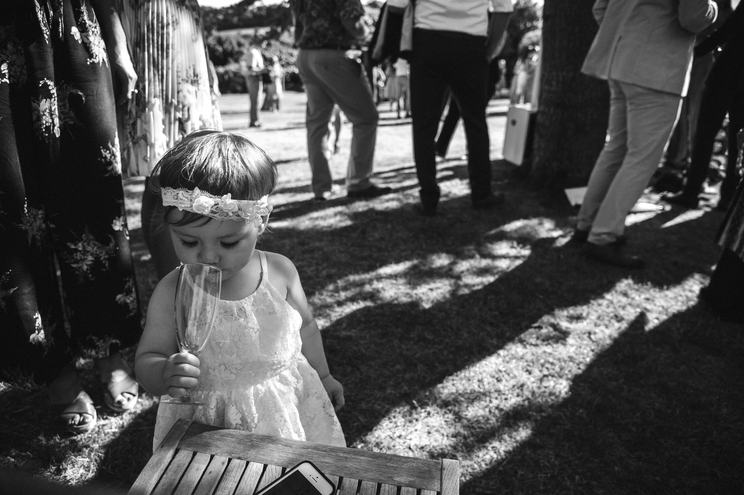A young girl in a lace dress and floral headband holding a glass, outdoors at a gathering with people in the background.